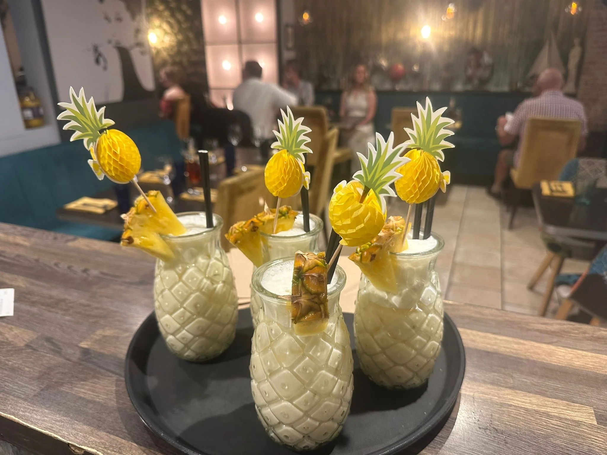 Several tropical drinks topped with paper pineapple decorations, served in pineapple-shaped glasses on a black tray in a restaurant setting.