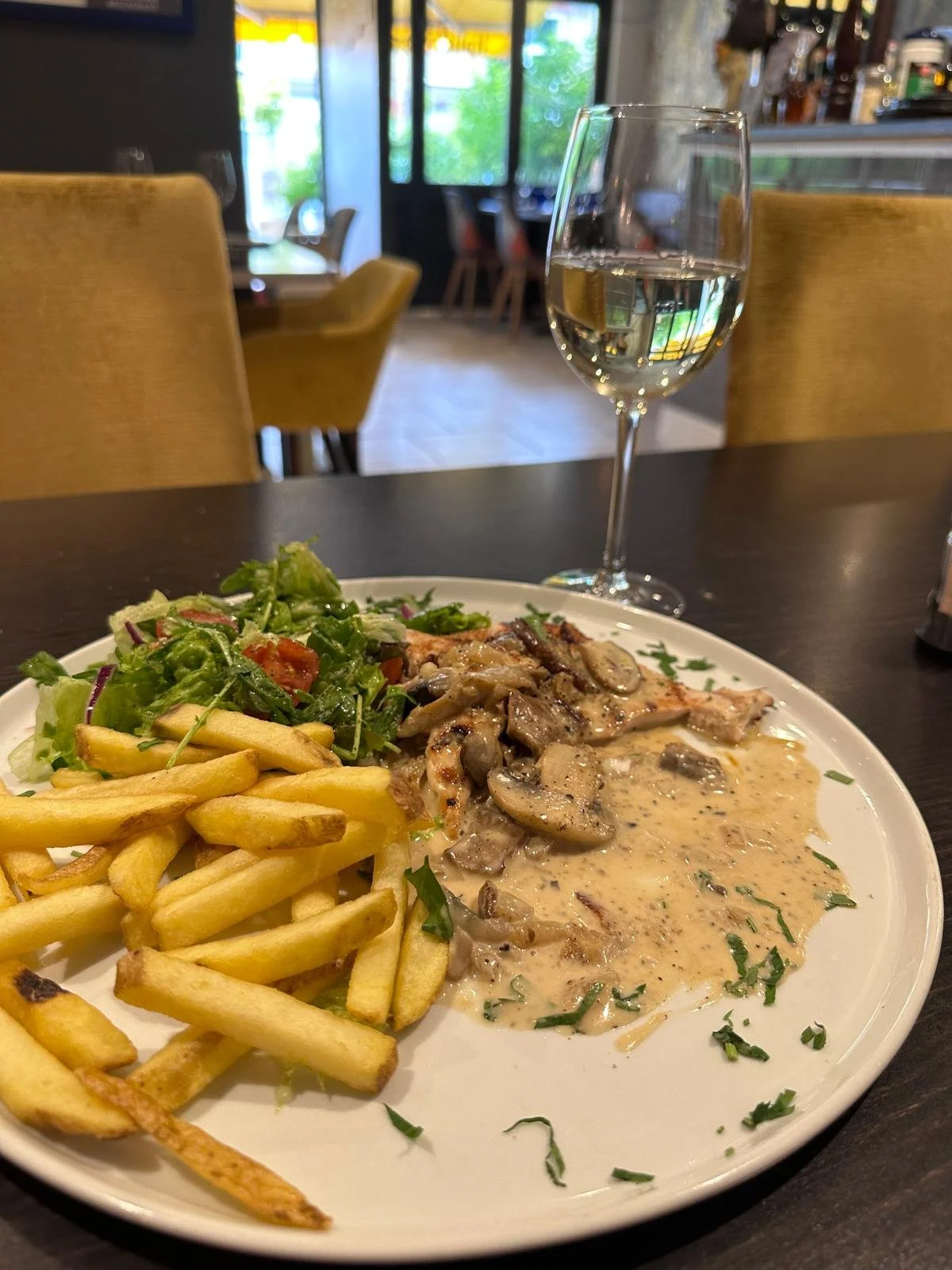 A plate of food including French fries, a mixed salad, and a chicken dish with mushroom gravy, with a glass of white wine on a dark table inside a restaurant.