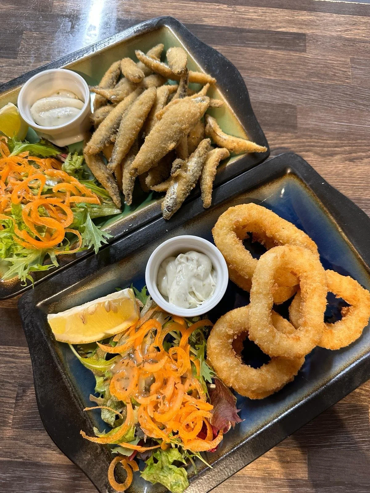 Two black rectangular plates with fried fish, onion rings, and salad with lettuce, orange slices, and shredded carrots, served with condiments in small white cups on a wooden table.
