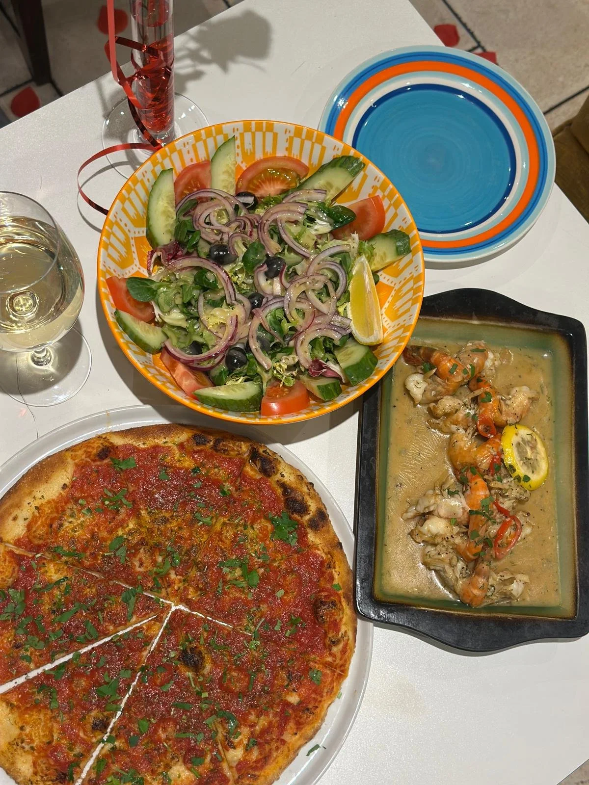 A table with a pizza, a salad in an orange bowl, a seafood dish in a black tray, a glass of white wine, a glass of water, and a blue and orange plate stack.