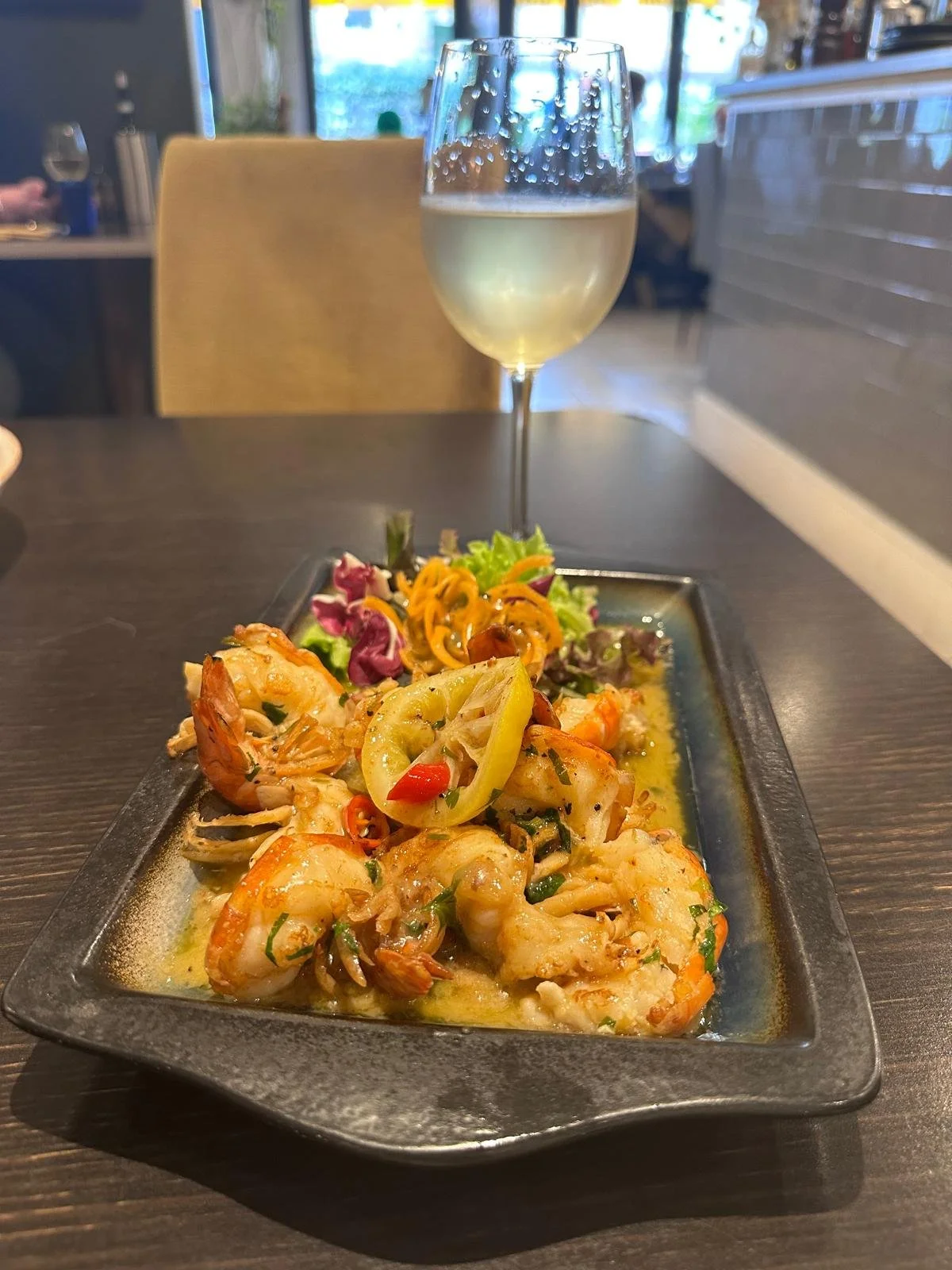 A black rectangular plate with cooked shrimp, sliced yellow peppers, and herbs. Behind the plate, there is a small salad with mixed greens and shredded carrots. A glass of white wine sits above the plate, with condensation on the glass. The background shows a restaurant setting with a wooden chair and a window.