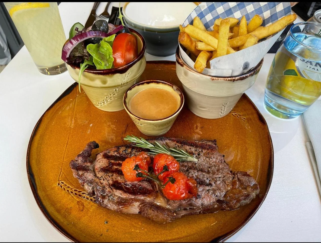 A plate with a cooked steak topped with cherry tomatoes and herbs, a small bowl of sauce, a side salad, a bowl of French fries, a glass of lemon water, and a drink in a tall glass on a white table.