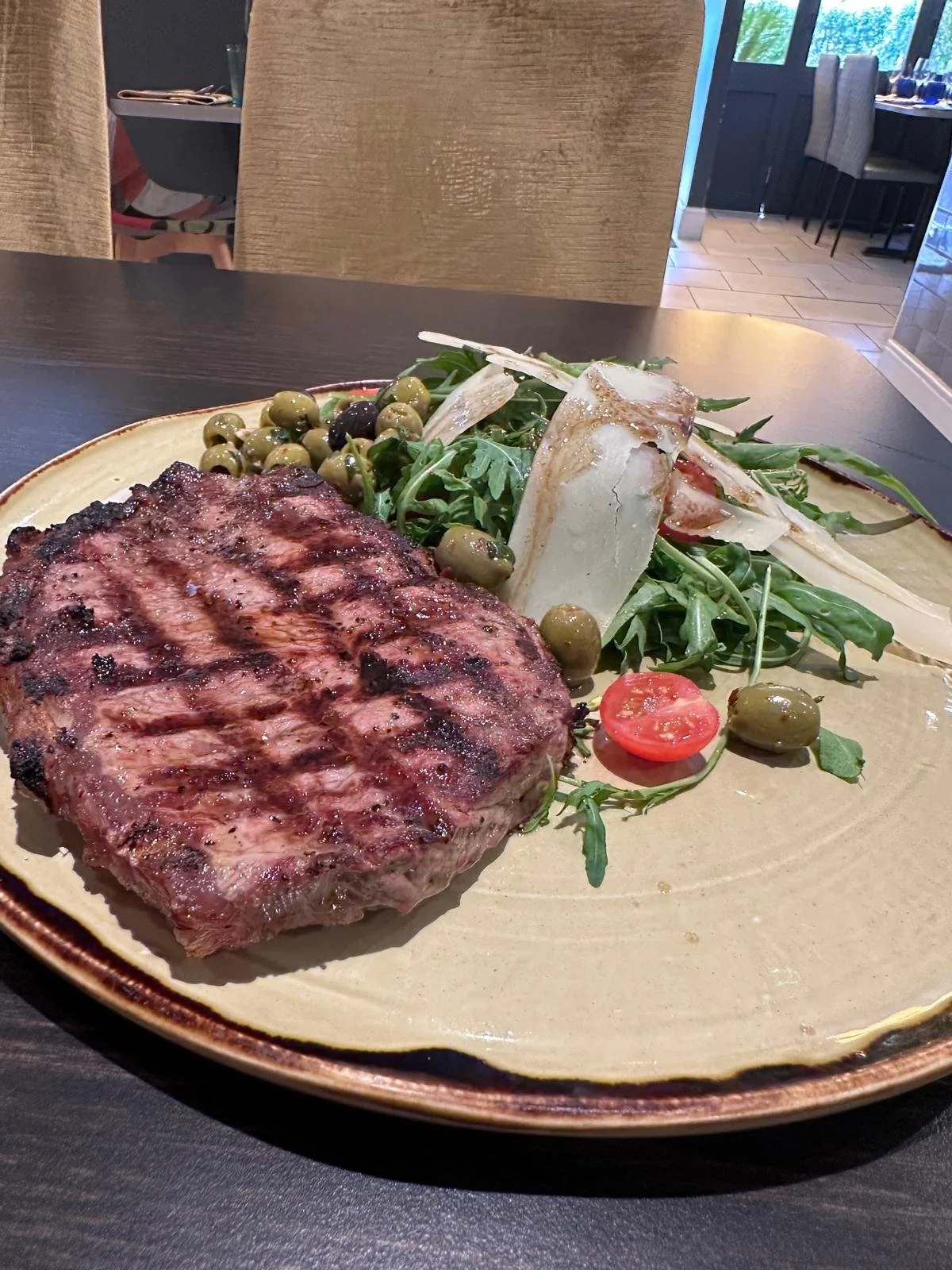 A plate with a grilled steak, cherry tomato slices, green olives, arugula, and cheese shavings, set on a dark dining table in a restaurant.