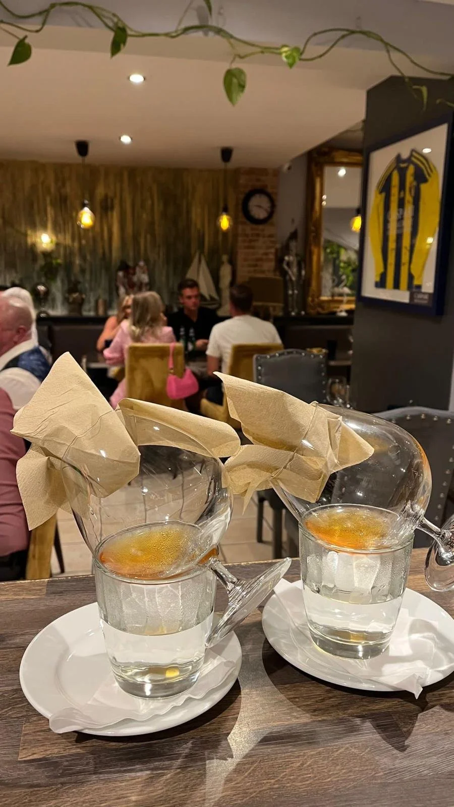 Two glasses of white wine with paper napkins inserted into the glasses, placed on white plates on a wooden table in a restaurant setting.