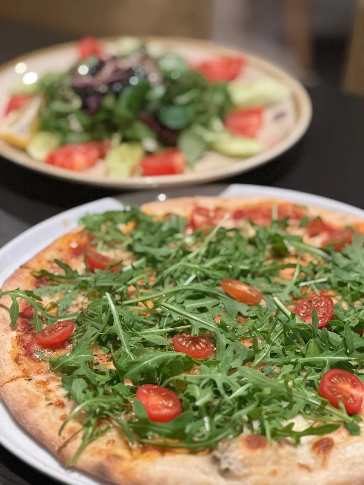 Close-up of a arugula and cherry tomato pizza on a white plate, with a salad in the background.