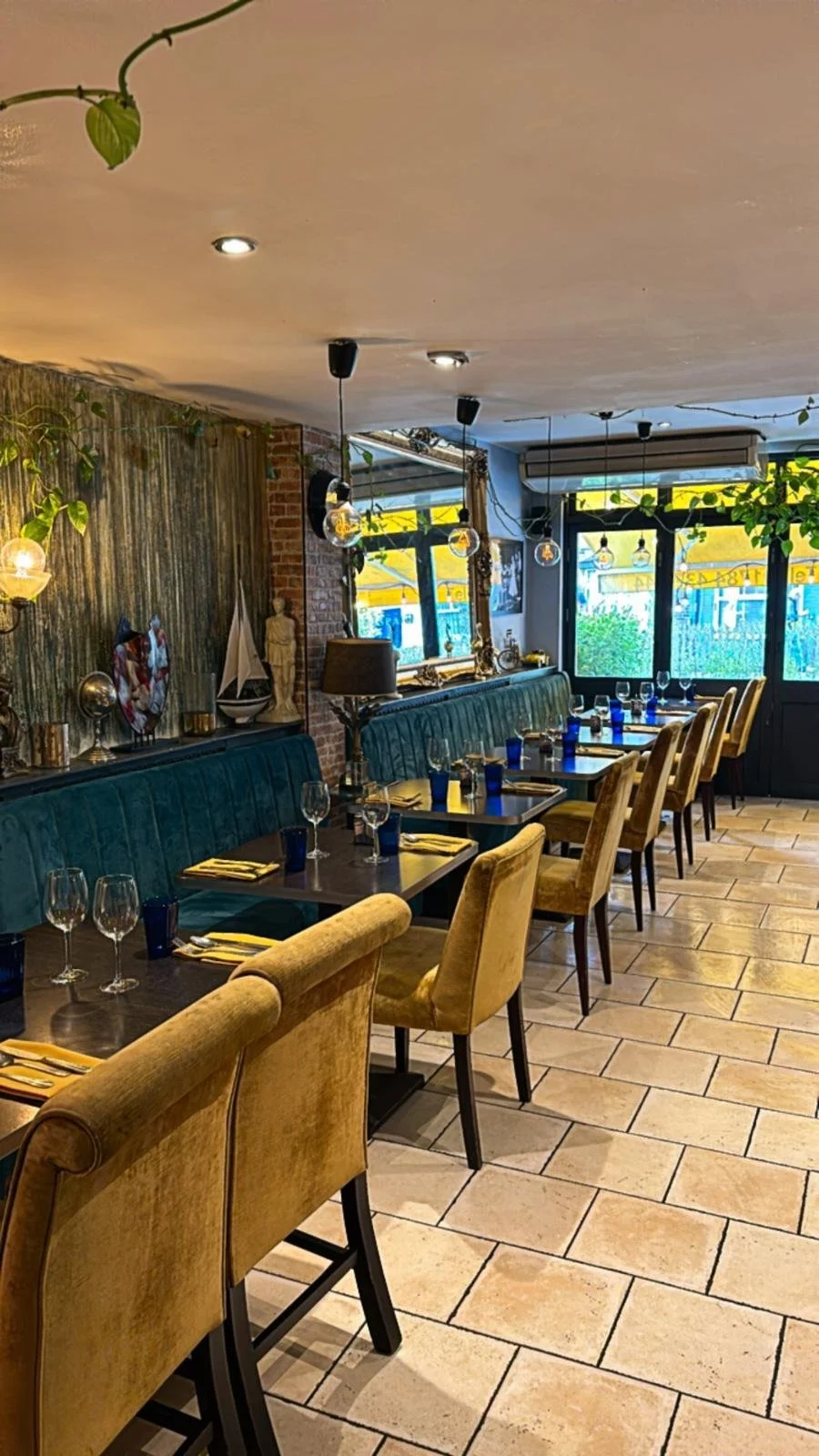 A cozy restaurant with a row of tables set for dining, featuring glasses, blue napkins, and yellow cloths. The space has a mix of yellow and teal upholstered chairs, a brick wall decorated with art and statues, warm lighting, and large windows with yellow awnings outside.