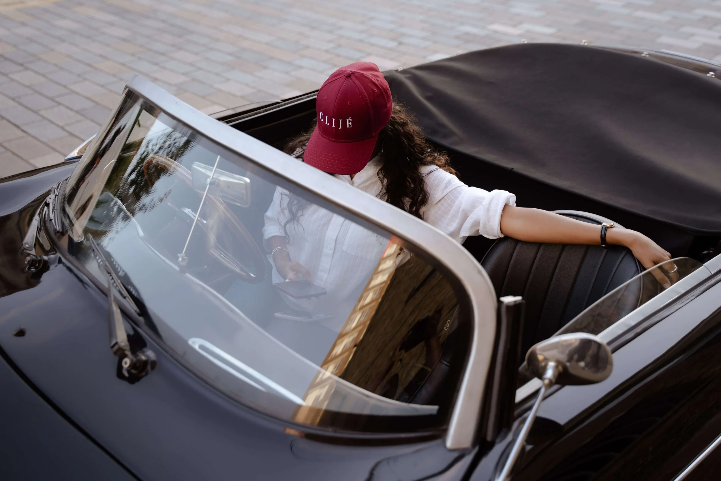 Woman with curly hair wearing a red baseball cap with 'CLIJÉ' written on it, sitting in a classic convertible car, on a paved street.