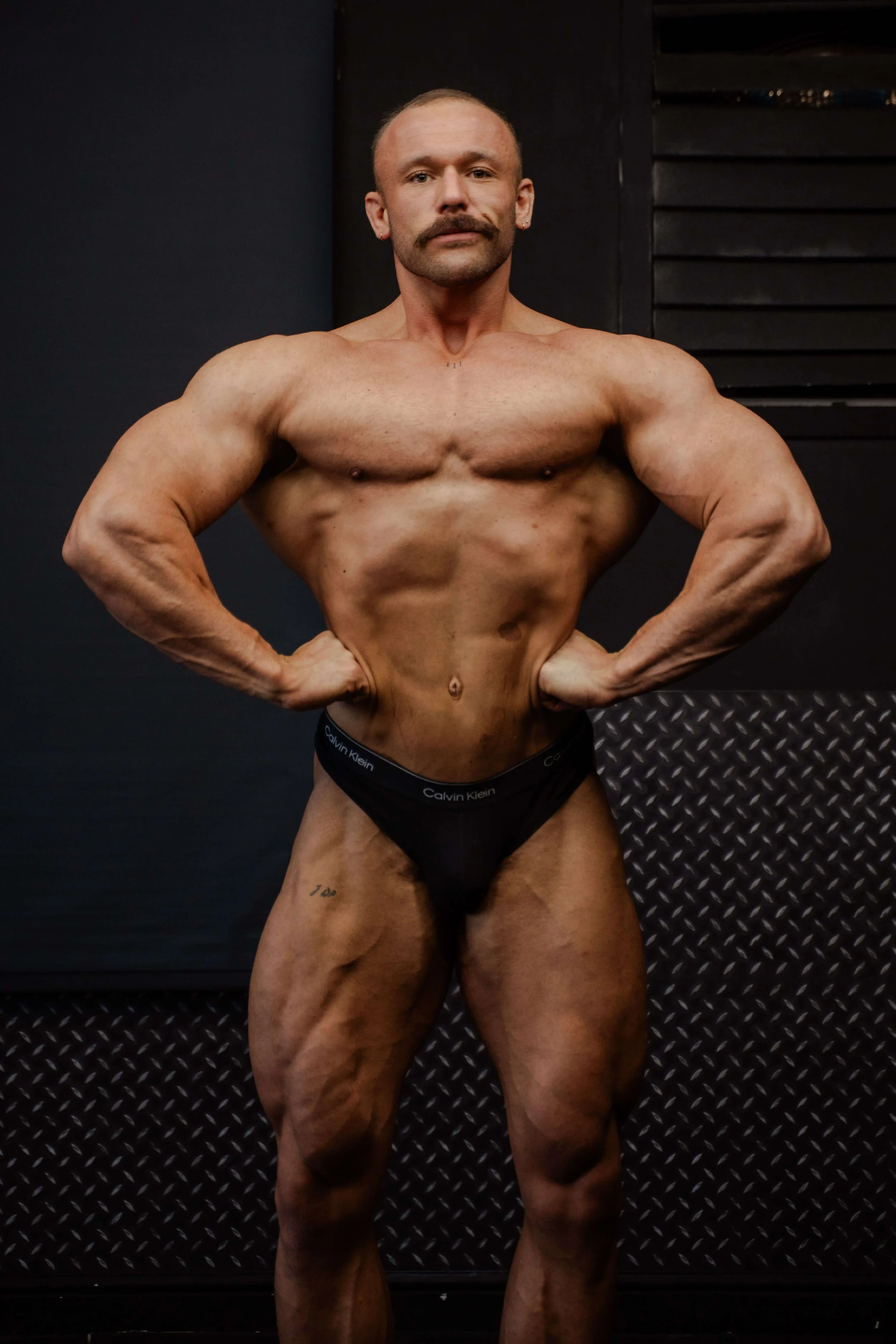 A muscular man with a mustache and tattoos posing with hands on hips in a gym.