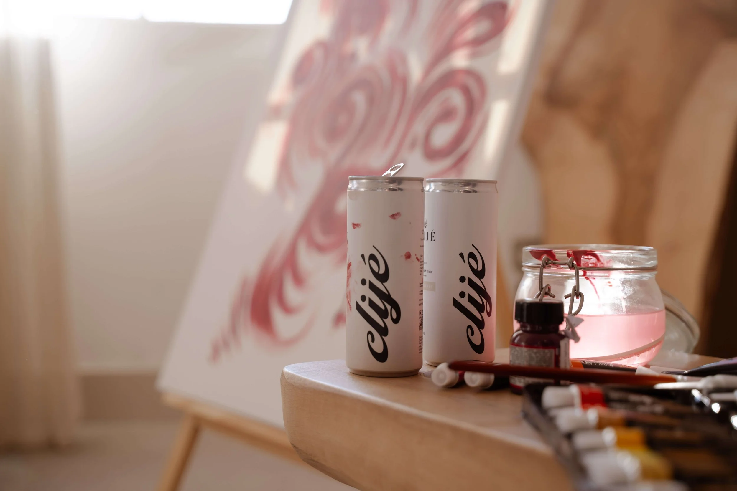 Two white cans of a beverage labeled 'Chisie' on a wooden table, next to glass jars with pink liquid, a small bottle, and some markers, with a blurred background of a wall with pink swirl artwork.