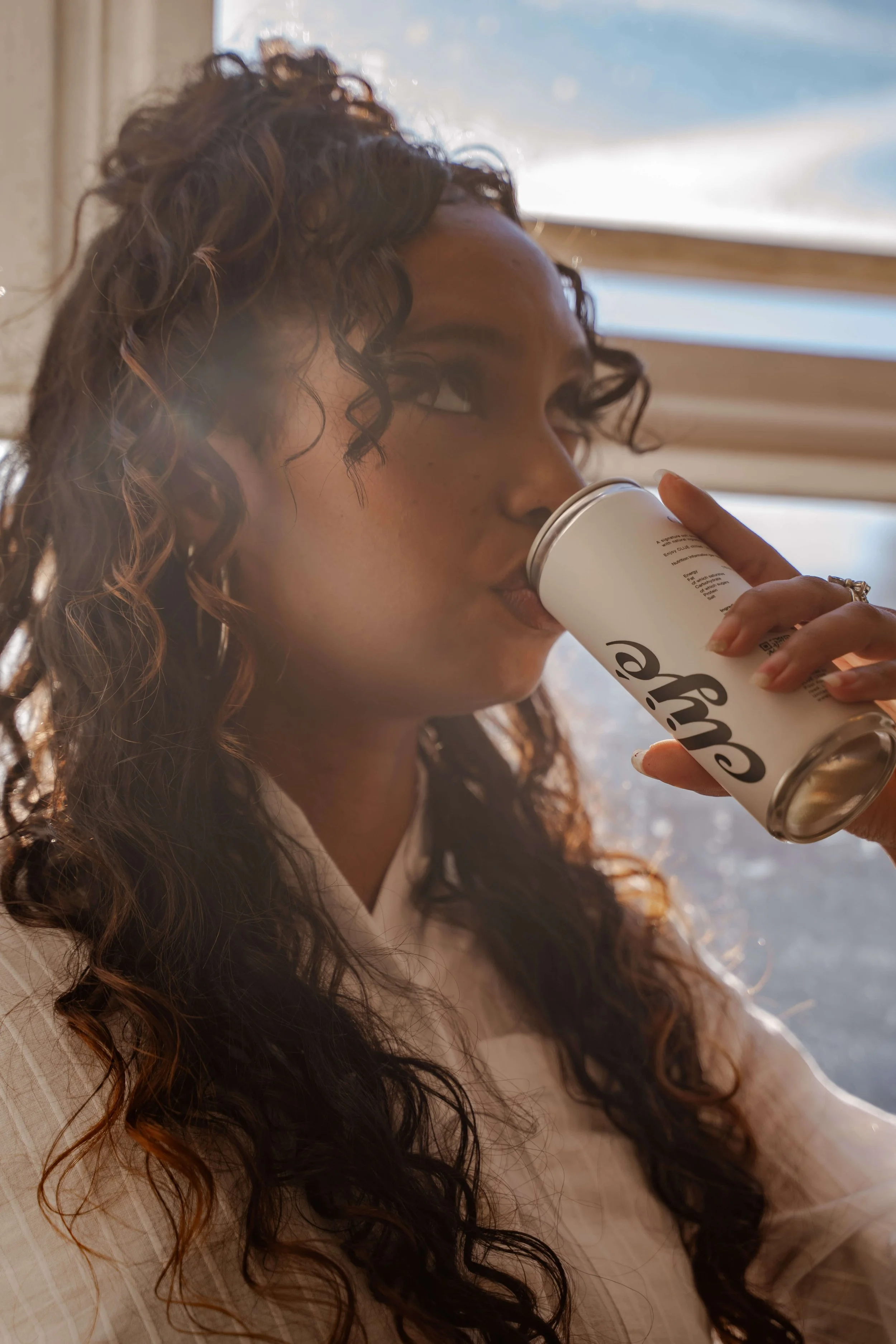 A woman with long curly hair is drinking from a white disposable coffee cup.