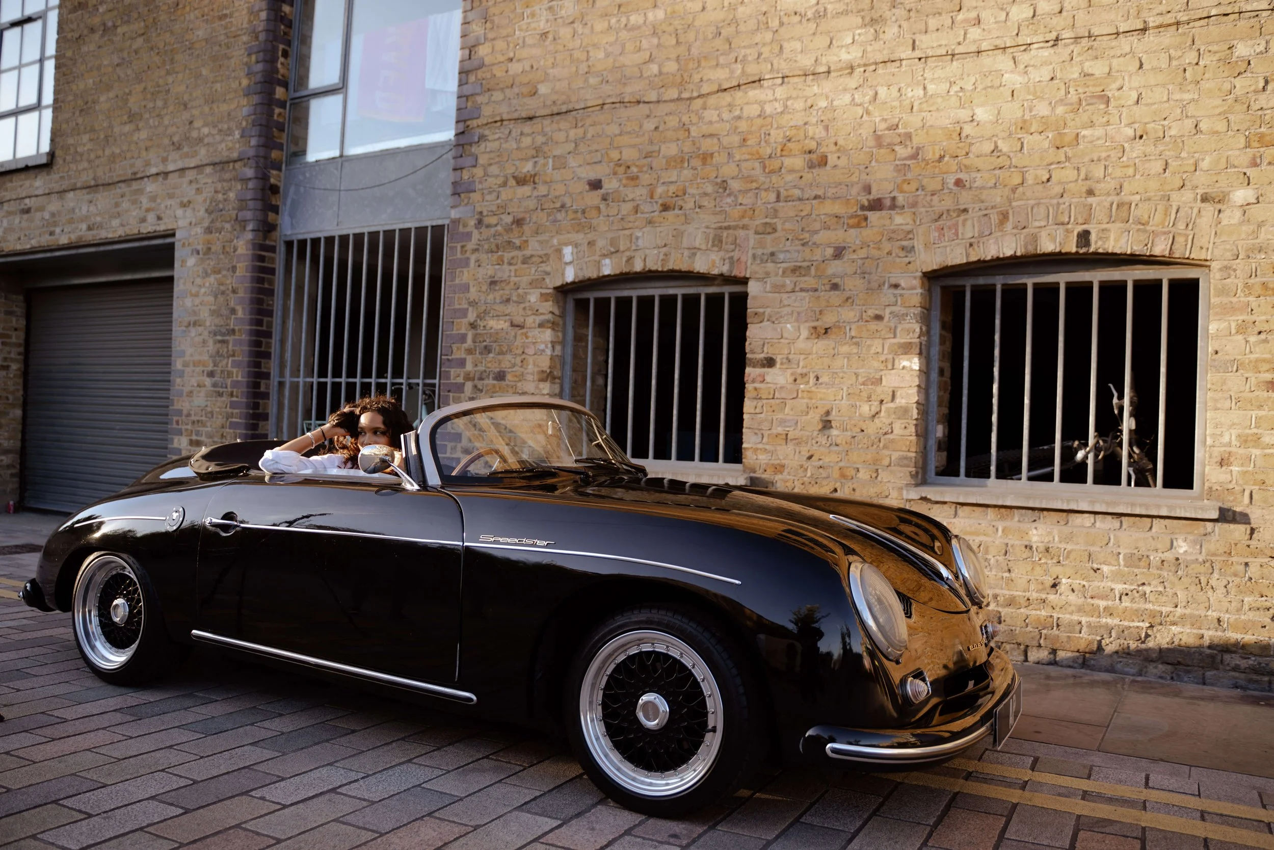 A woman with long hair sitting in a black vintage convertible sports car parked on a brick street in front of a brick building with barred windows.