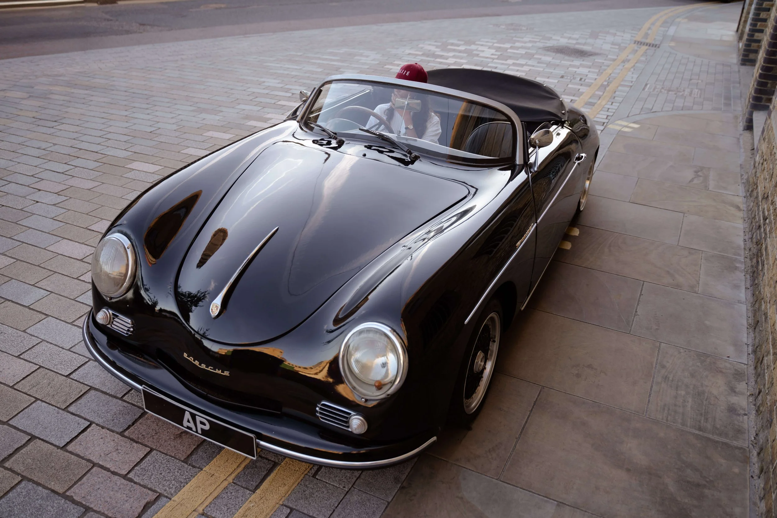 Black vintage Porsche convertible car parked on a street with two people inside, one woman taking a selfie with her phone, wearing a white blazer and red hat. The car is parked next to a brick wall and a paved sidewalk.