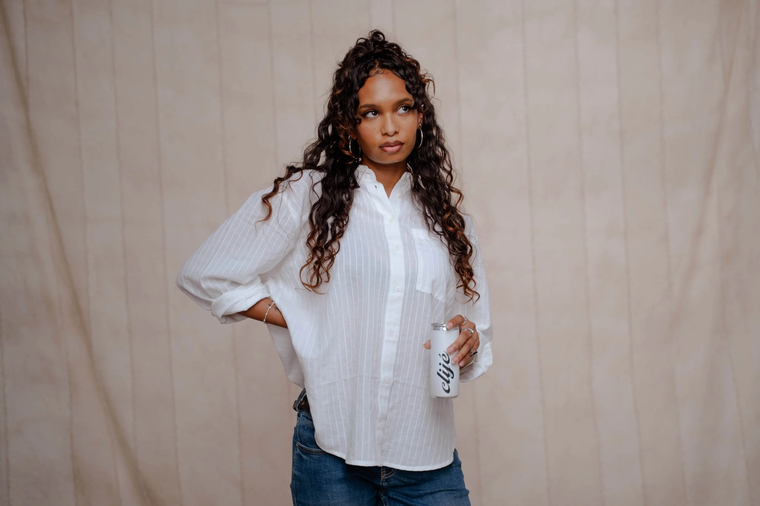 A woman with curly hair, wearing a white striped button-up shirt and blue jeans, stands with one hand on her hip, holding a white coffee tumbler with black lettering in her other hand, against a neutral background.