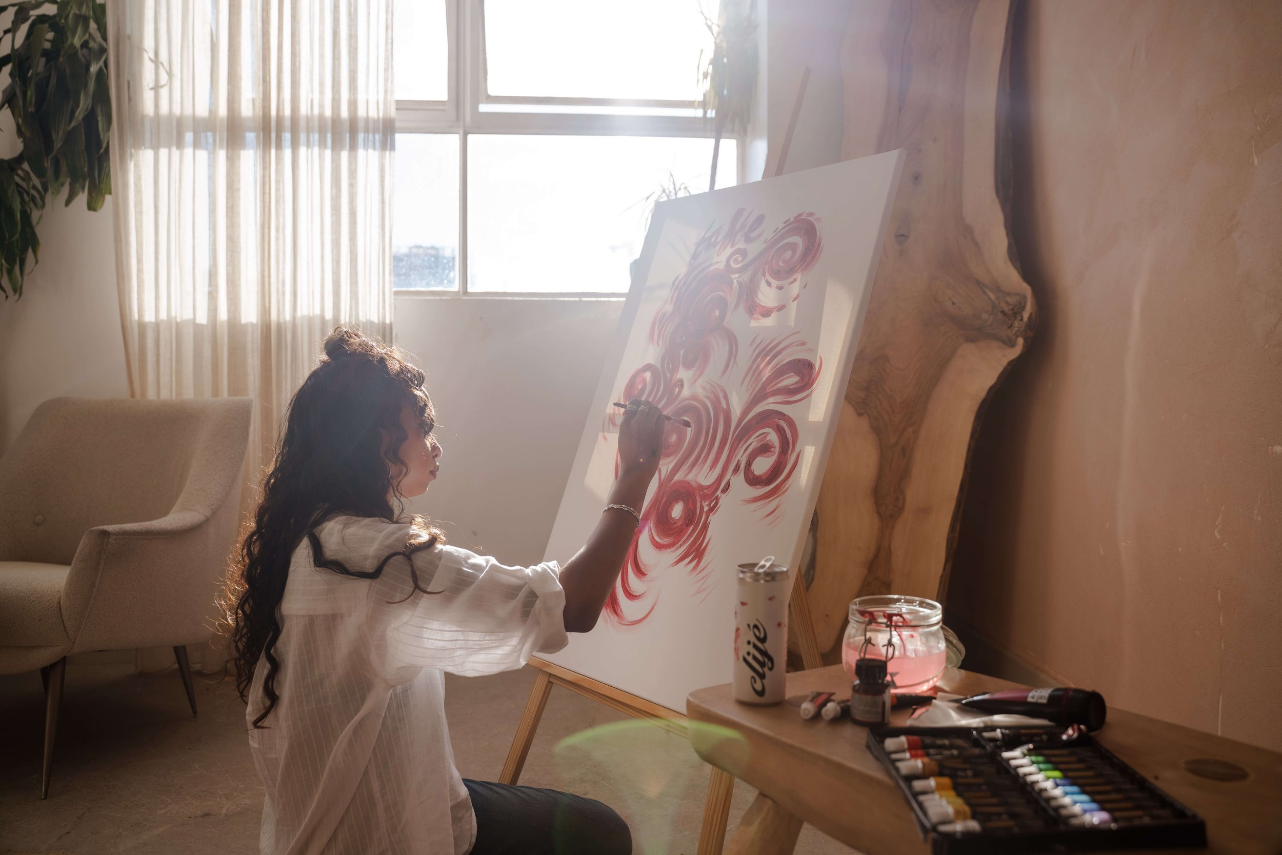 A woman with curly hair painting abstract swirls in red on a canvas, seated on the floor near a window with sheer curtains, with painting supplies and a jar of pink paint nearby.