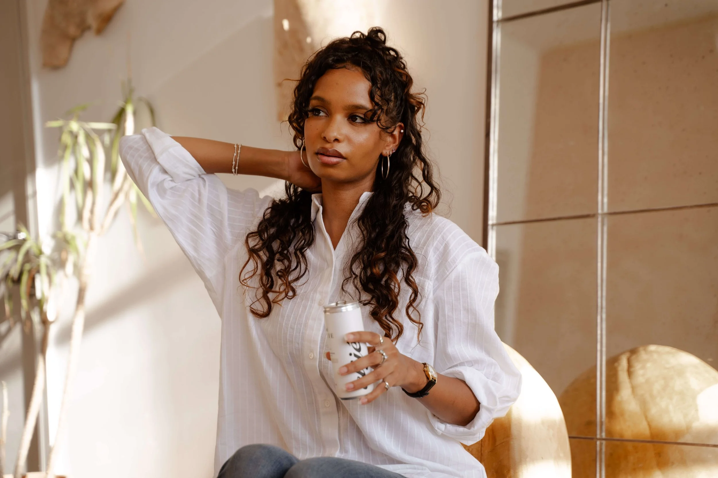 A woman with curly hair, earrings, and a watch, wearing a white shirt, standing indoors near a mirror, holding a drink, with a plant and beige tiled wall in the background.