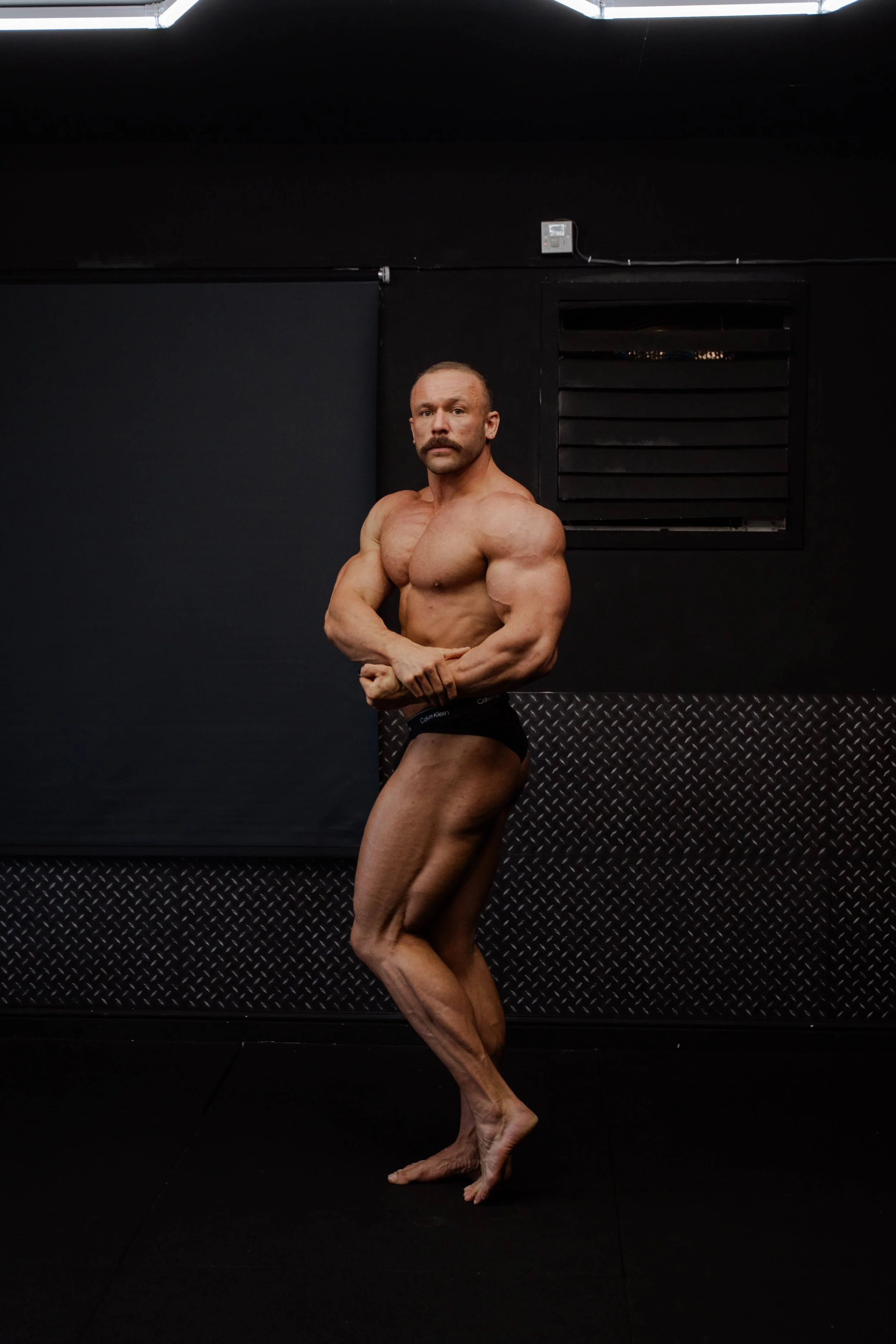 A muscular man with a mustache and no shirt, wearing black underwear, standing against a black background in a gym or workout space.