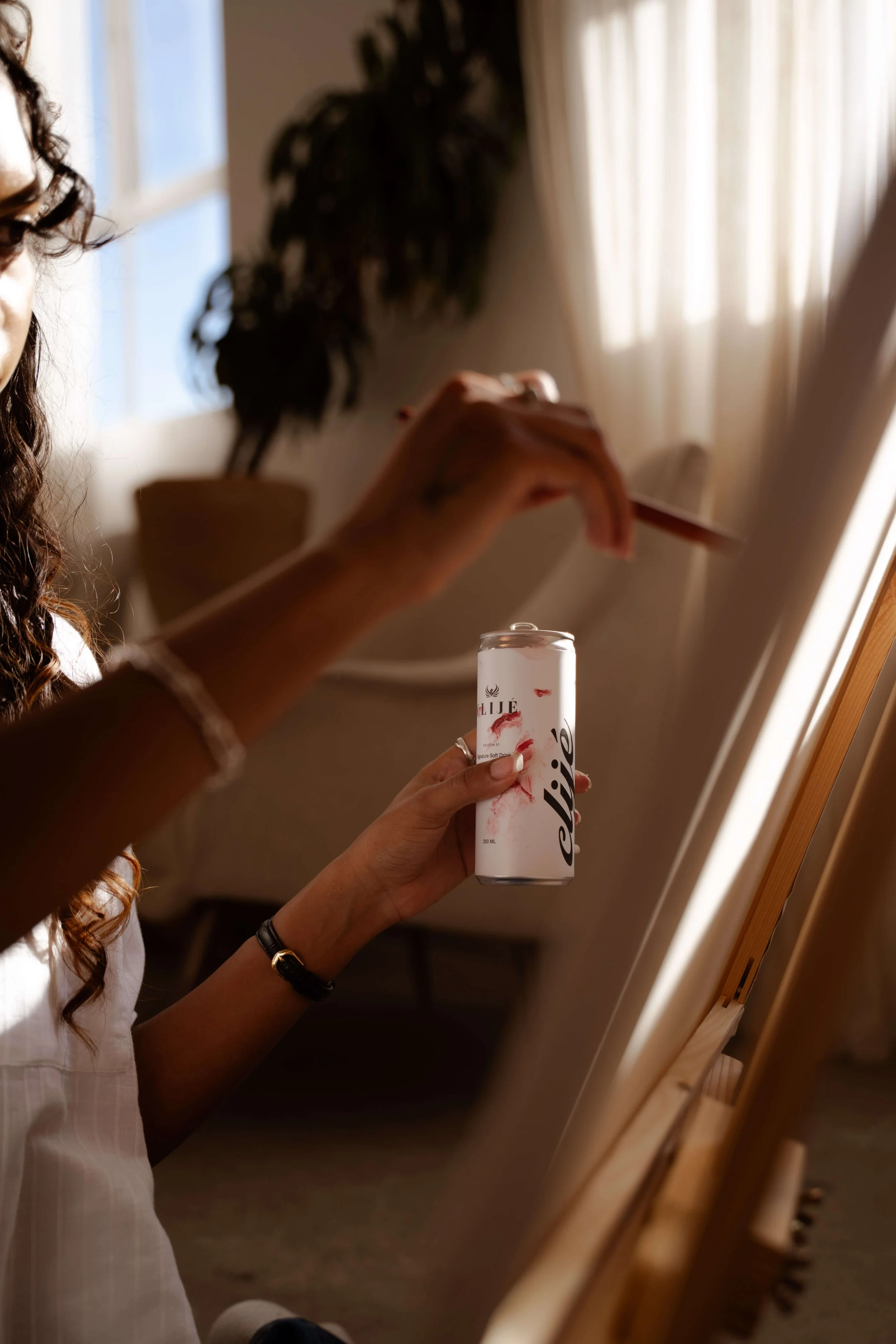 Person with curly brown hair wearing a white shirt, holding a white can of La Vie sparkling water, standing in front of a mirror with sunlight shining through curtains.