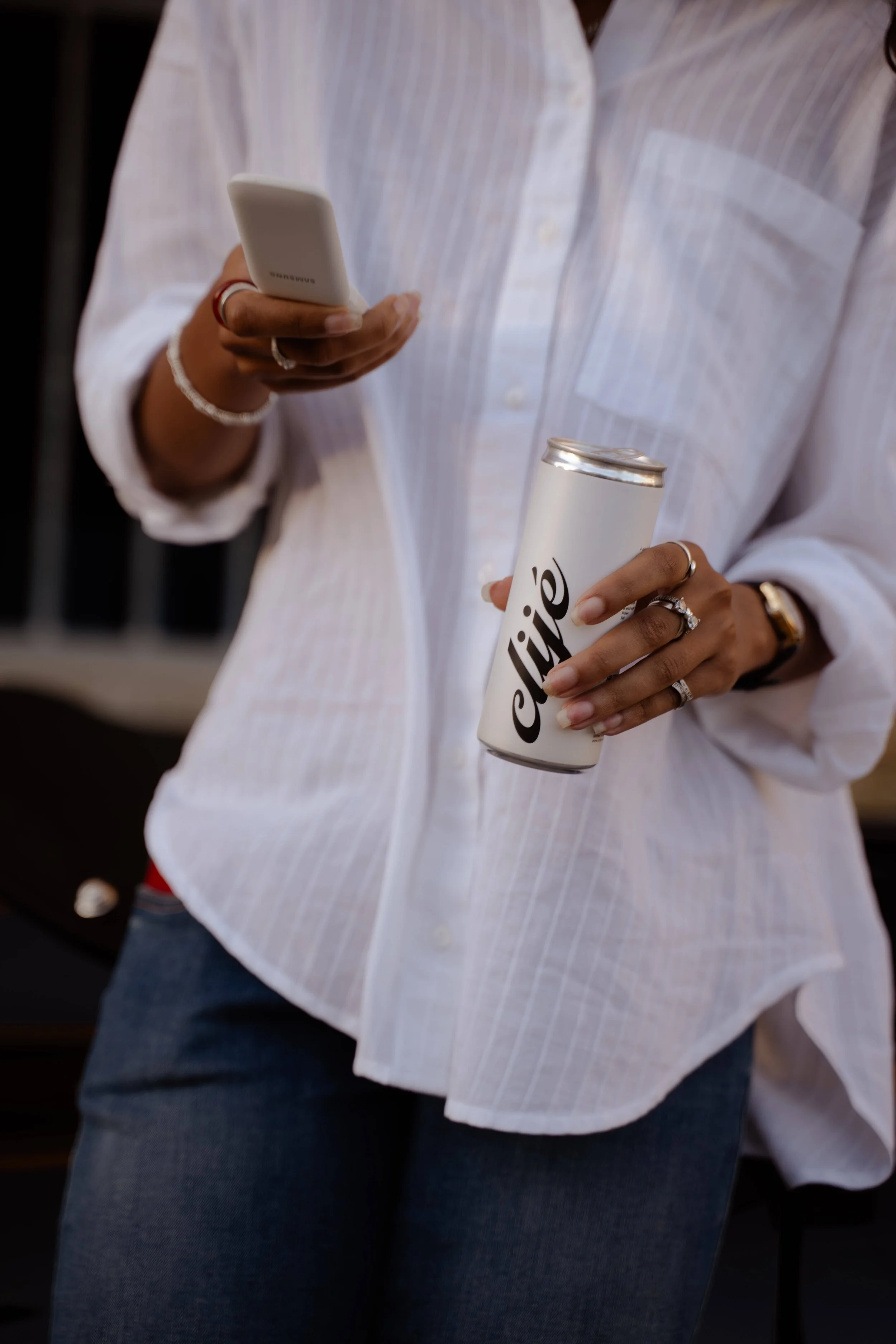 Person wearing a white striped shirt holding a white Samsung mobile phone and a canned beverage.