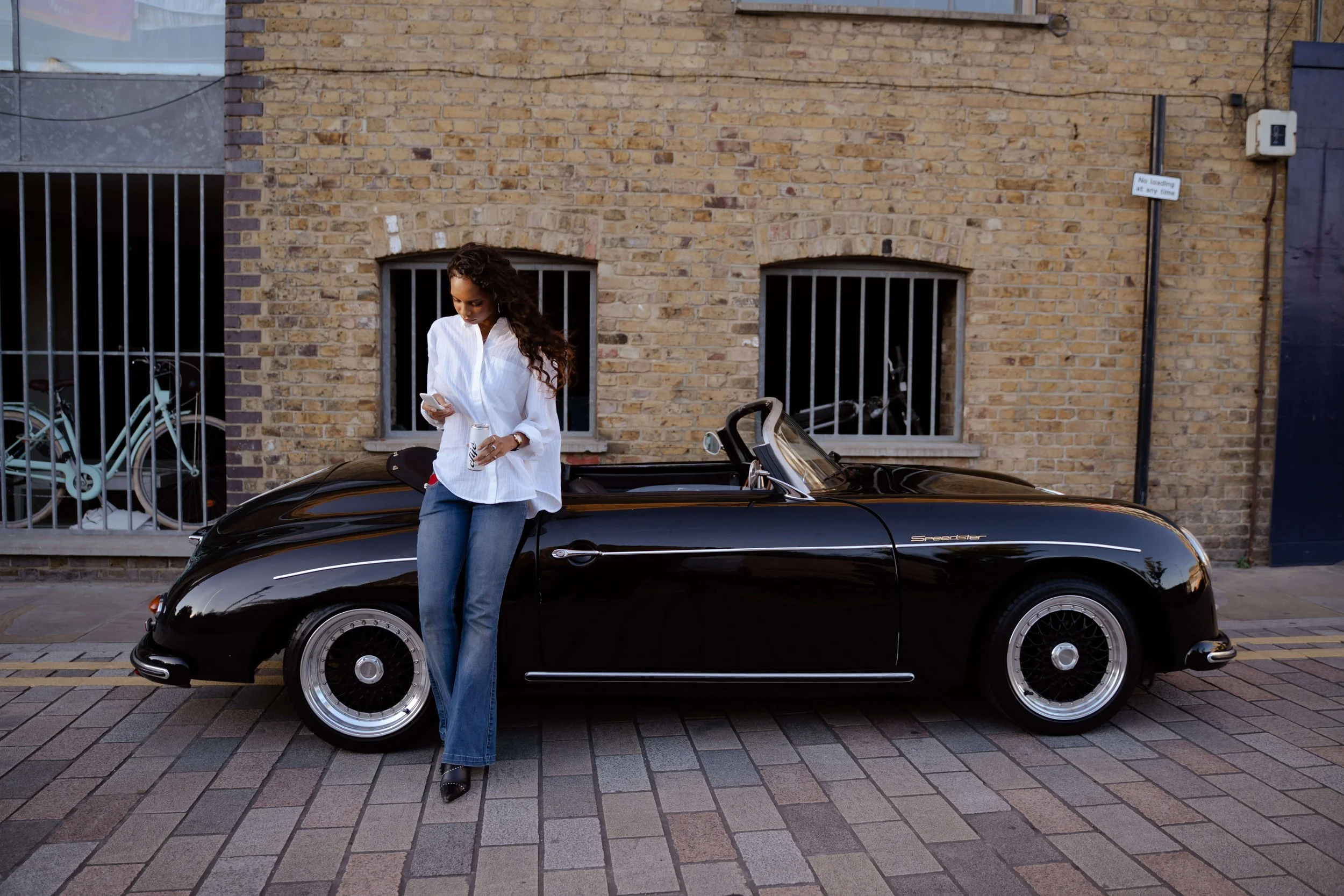 A woman in a white blouse and jeans standing next to a black sports car on a city street with a brick building in the background and a sign on the wall that says 'No loading at any time.'