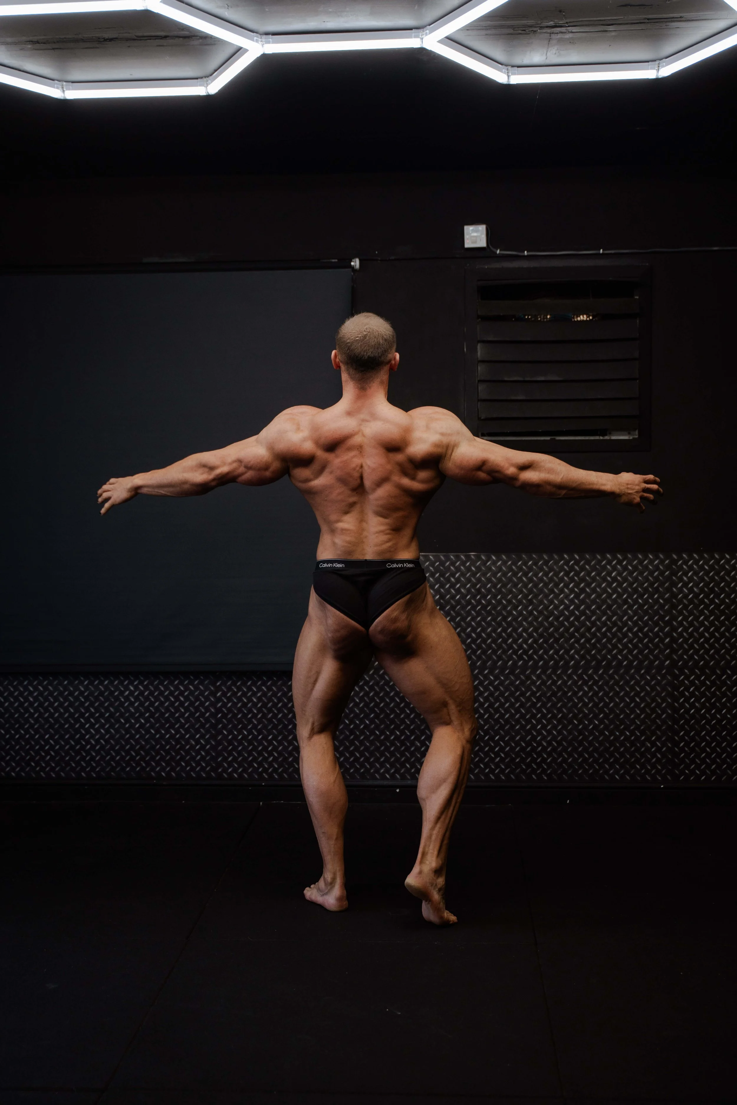 A muscular man with a shaved head performs a pose in a black gym with a low ceiling light fixture.