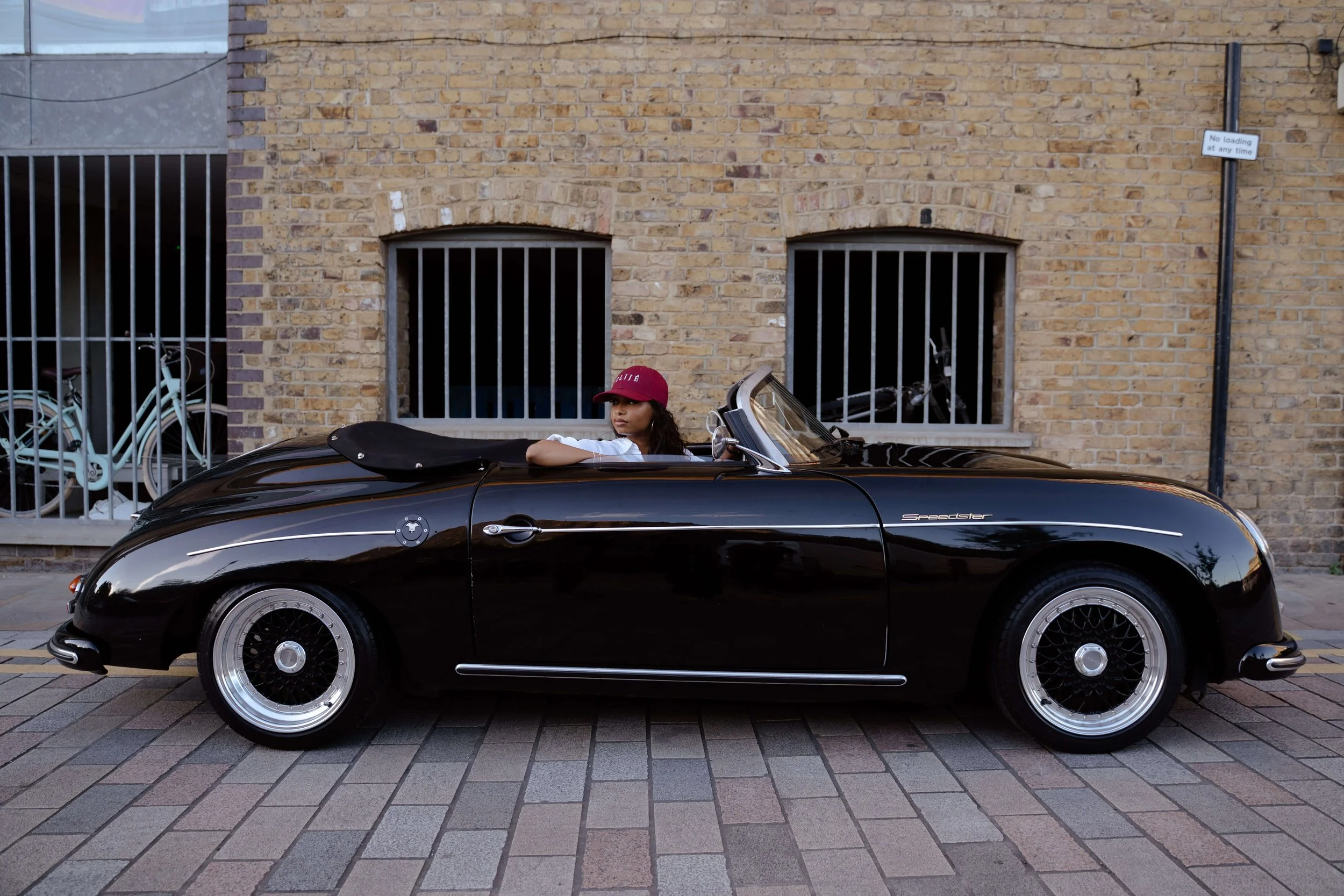 A young woman with curly hair, wearing a red cap and white shirt, sitting in a black vintage convertible car parked on a city street. The background features a brick wall with two barred windows and a bicycle behind a metal fence.