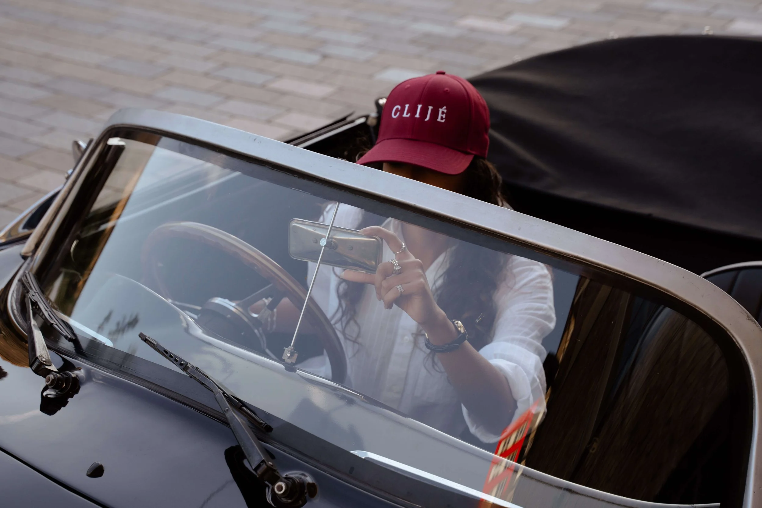 A woman wearing a red cap with the word 'CLIQUE' on it sits in a vintage convertible car, taking a selfie.