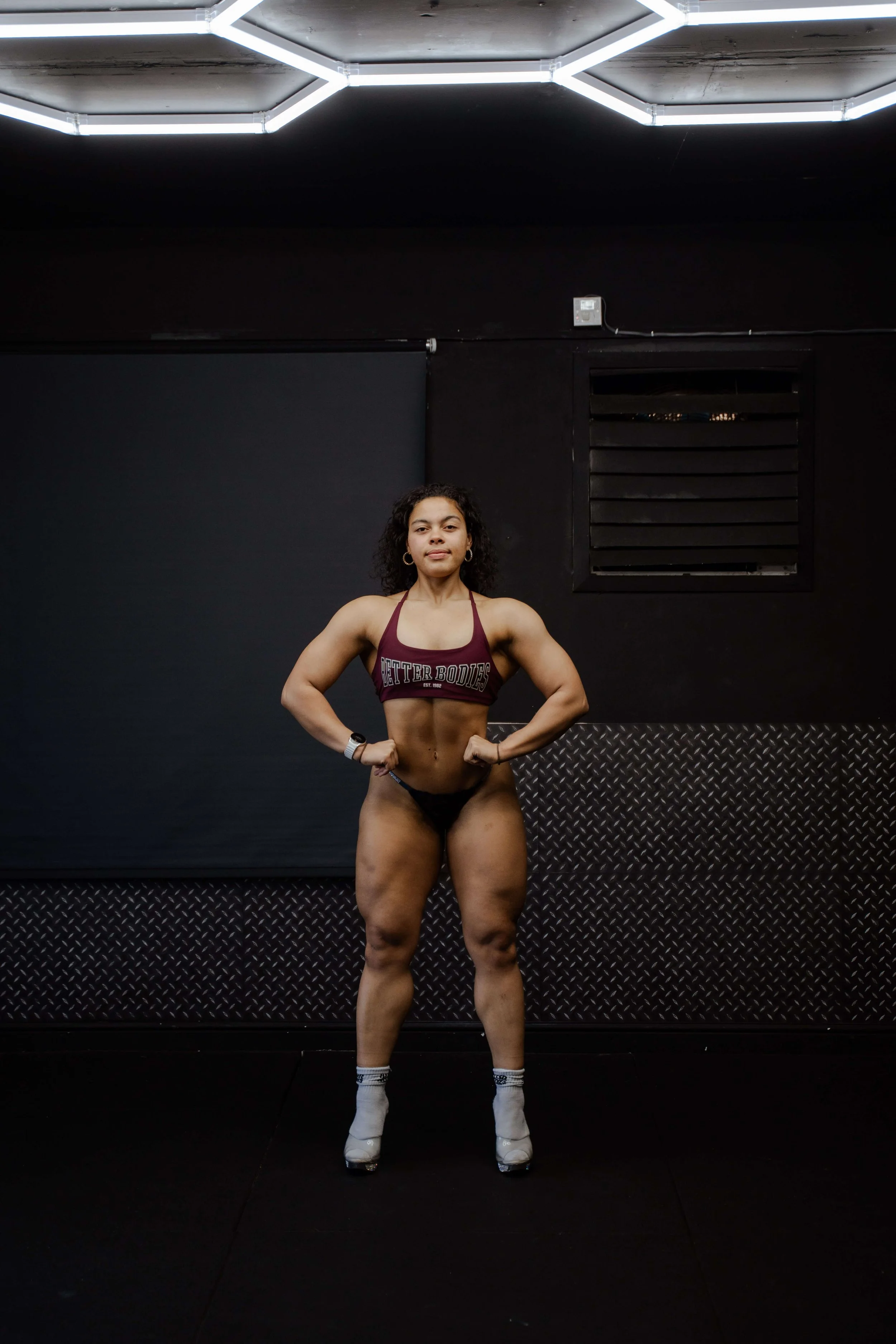 A woman standing confidently with hands on hips in a gym, wearing a maroon sports bra with 'Better Bodies' written on it, black shorts, white socks, and high-heeled shoes, against a dark wall with industrial lighting.