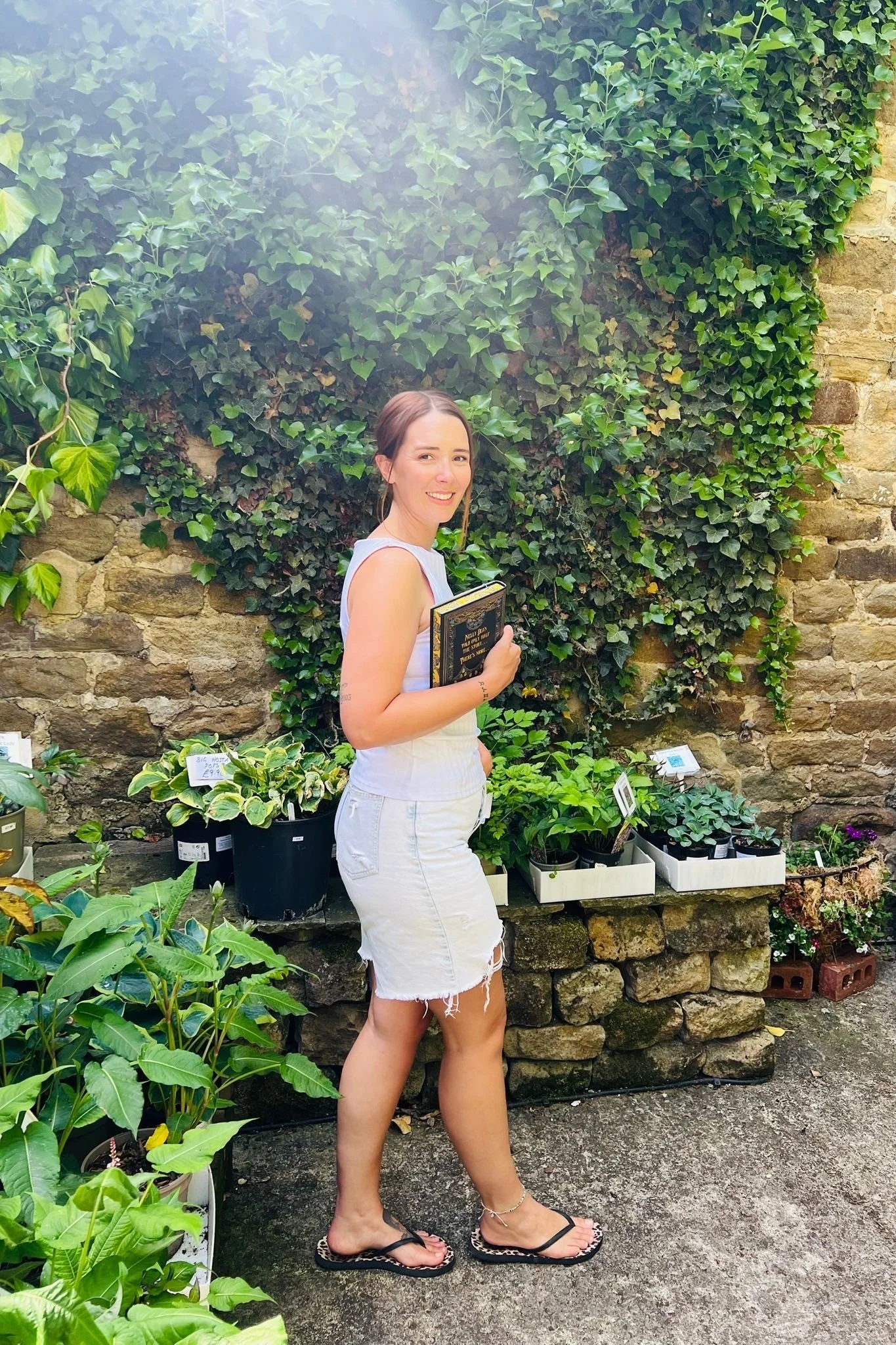 Author Amy Brook smiling at camera. She is holding a book and has plants in the background. She is wearing a white vest, jean shorts and flip flops.