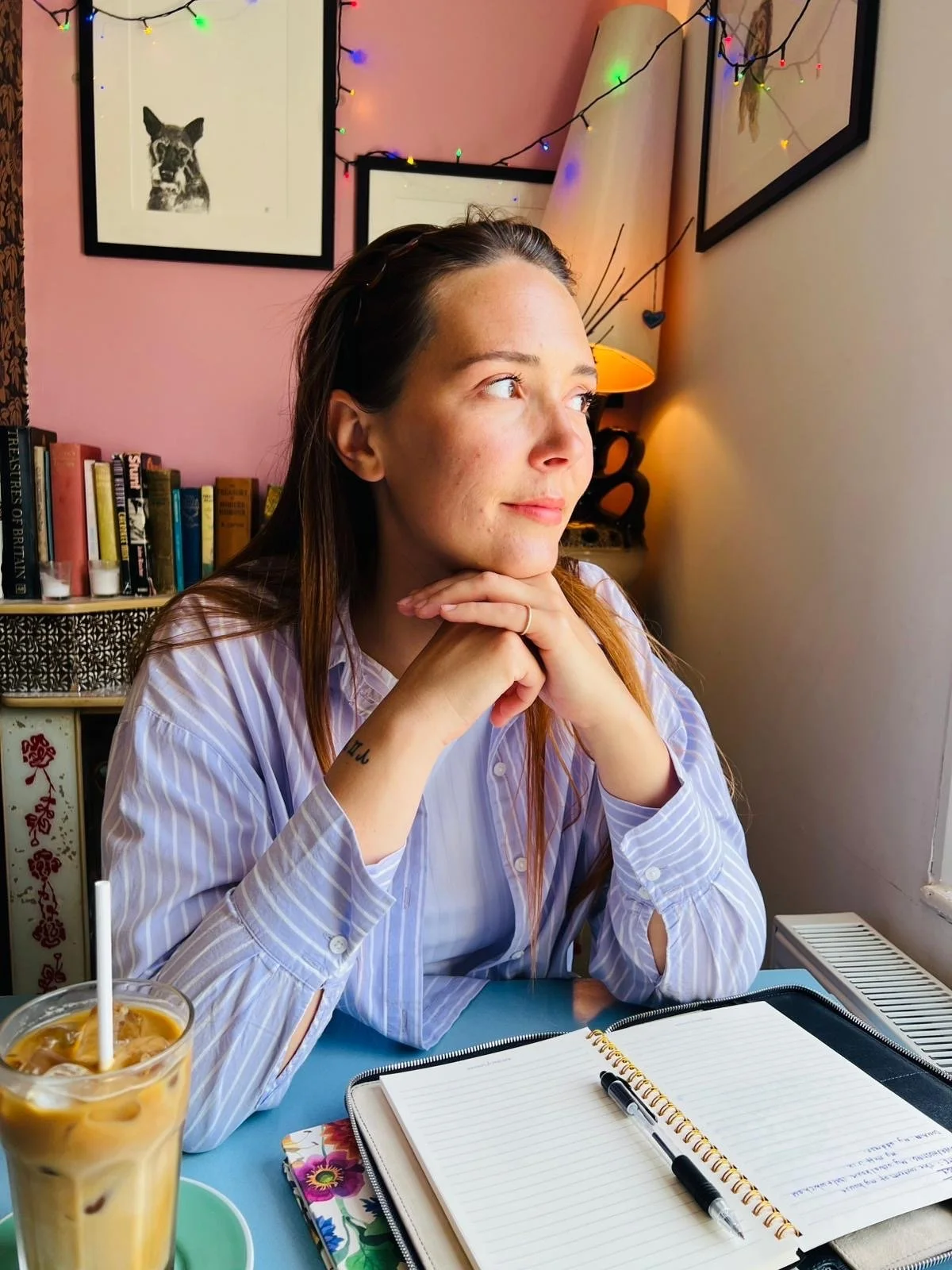 Author Amy Brook sitting in a cafe. She is wearing a lilac shirt with white stripes and looking out of the window. Iced coffee, notebook and pen on the table. Behind is a pink wall, books, colourful fairy lights and prints of dogs on the wall