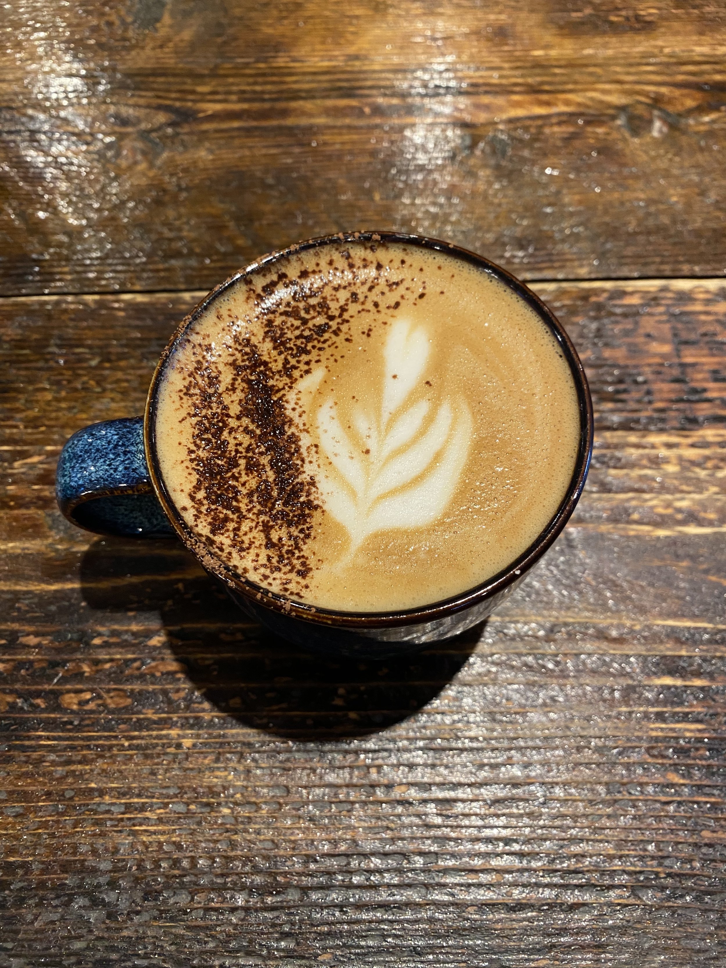 A cup of cappuccino with coffee art and chocolate sprinkles in a blue artisan mug on a wooden table.