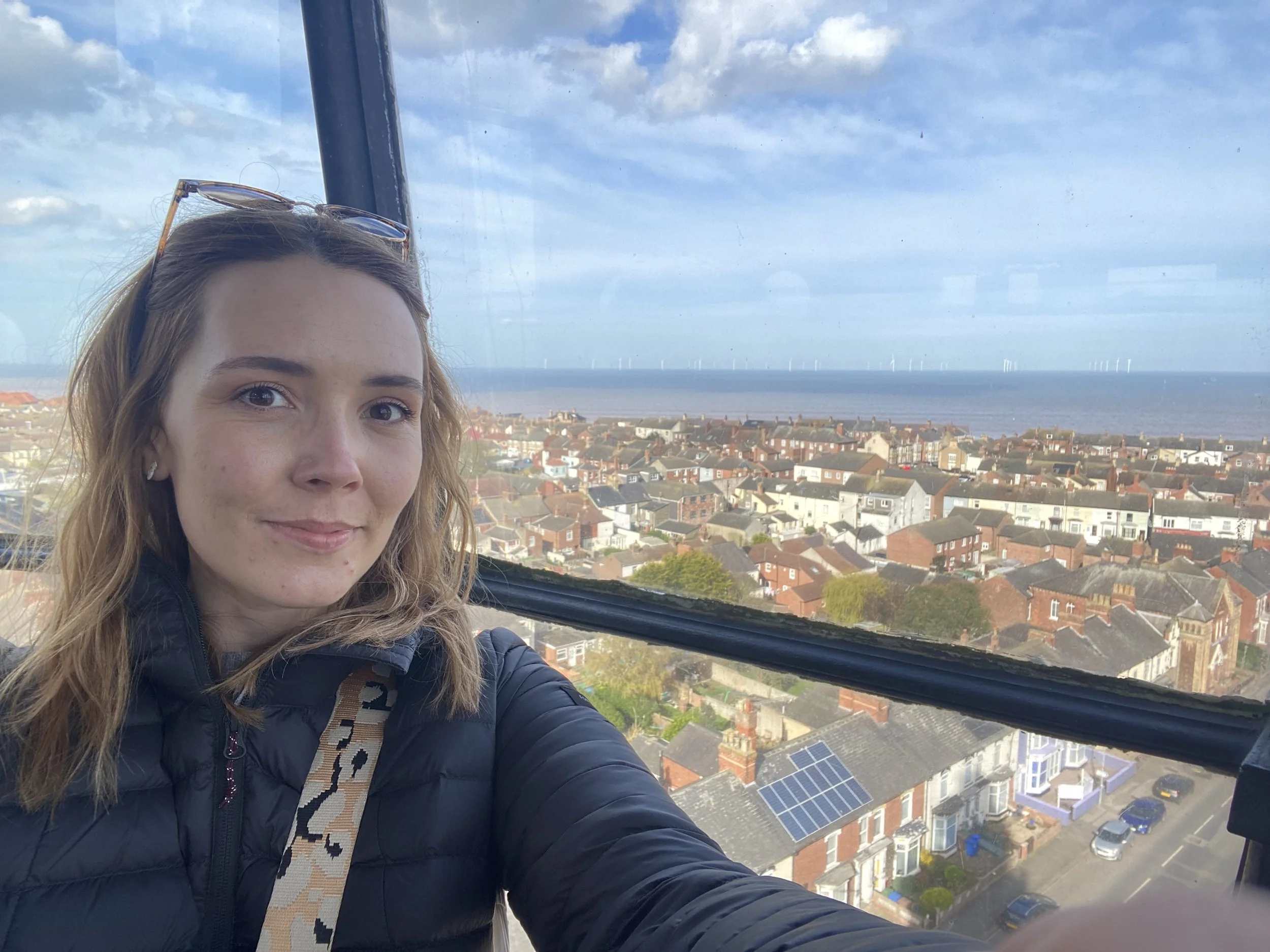Selfie of author Amy Brook at top of a lighthouse on Yorkshire East Coast. Houses and the sea can be seen in the background. She is wearing a black coat, leopard print bag strap and sunglasses on her head.