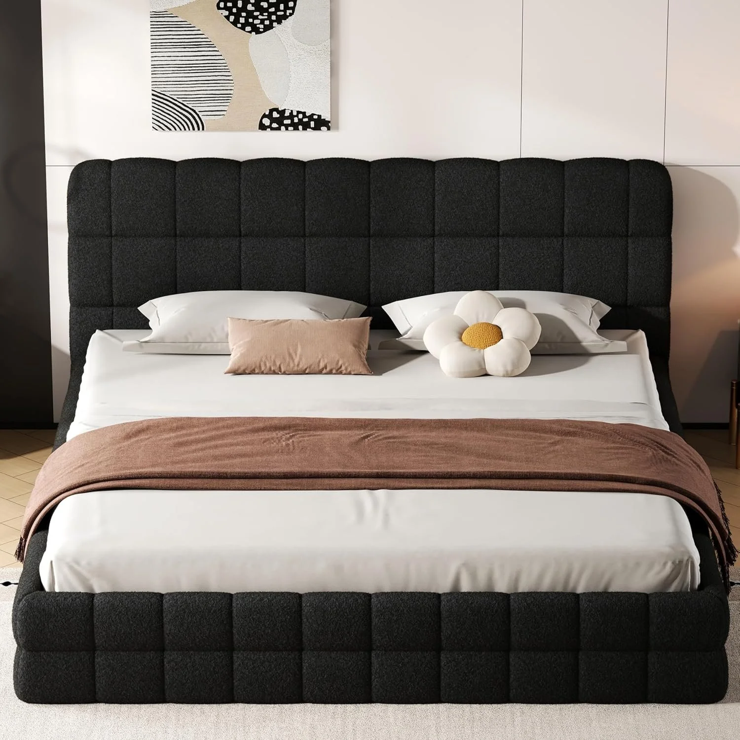 A modern bedroom with a dark upholstered bed, white bedsheets, a pink pillow, a brown blanket, and a flower-shaped pillow. There's a piece of abstract black and white wall art above the bed.