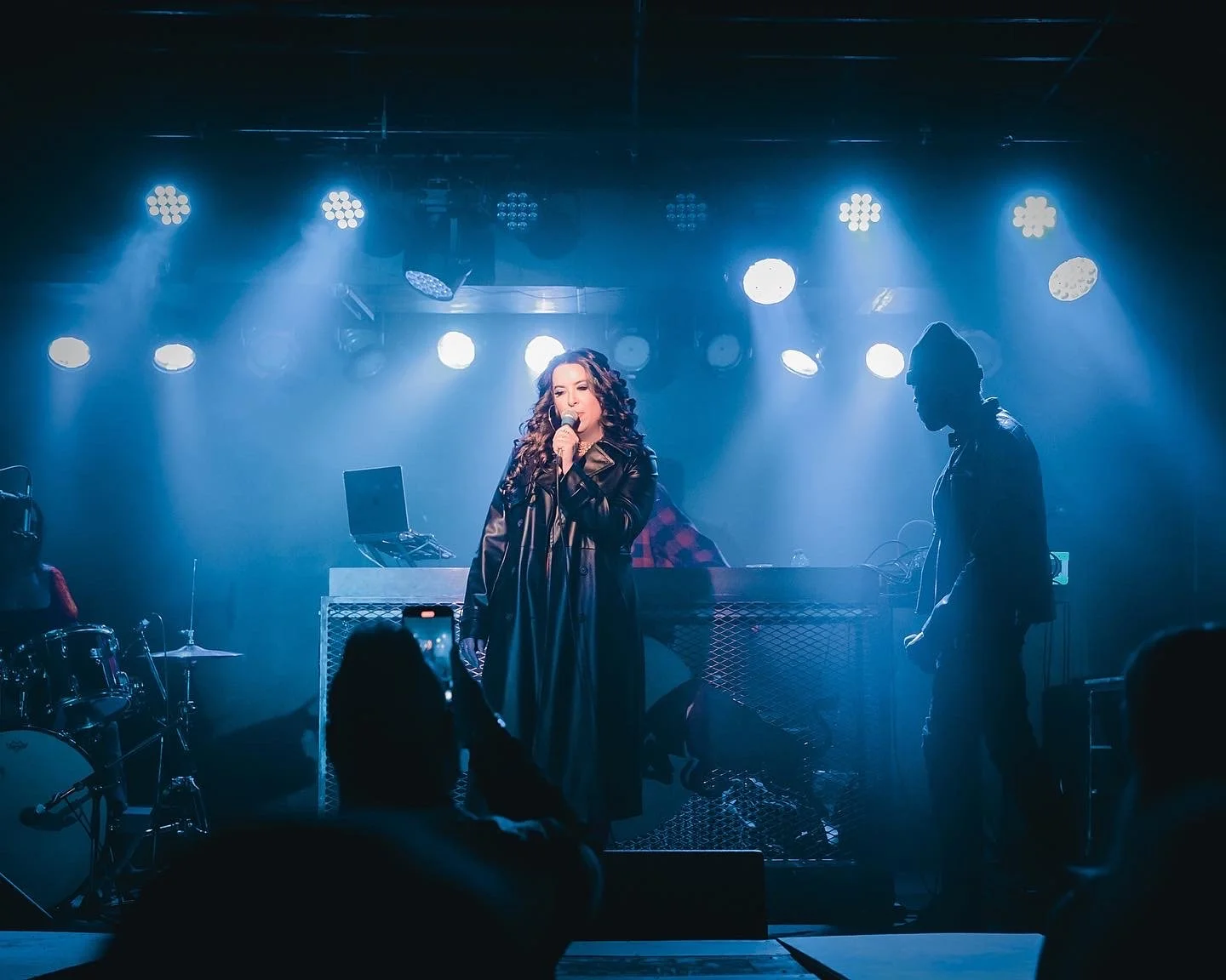 Kelly Besd, artist, with curly hair performs on stage with a microphone, accompanied by ChefBeatz and L.I Freeze. The stage is illuminated with bright white and blue lights, with musical instruments like drums visible on the side.