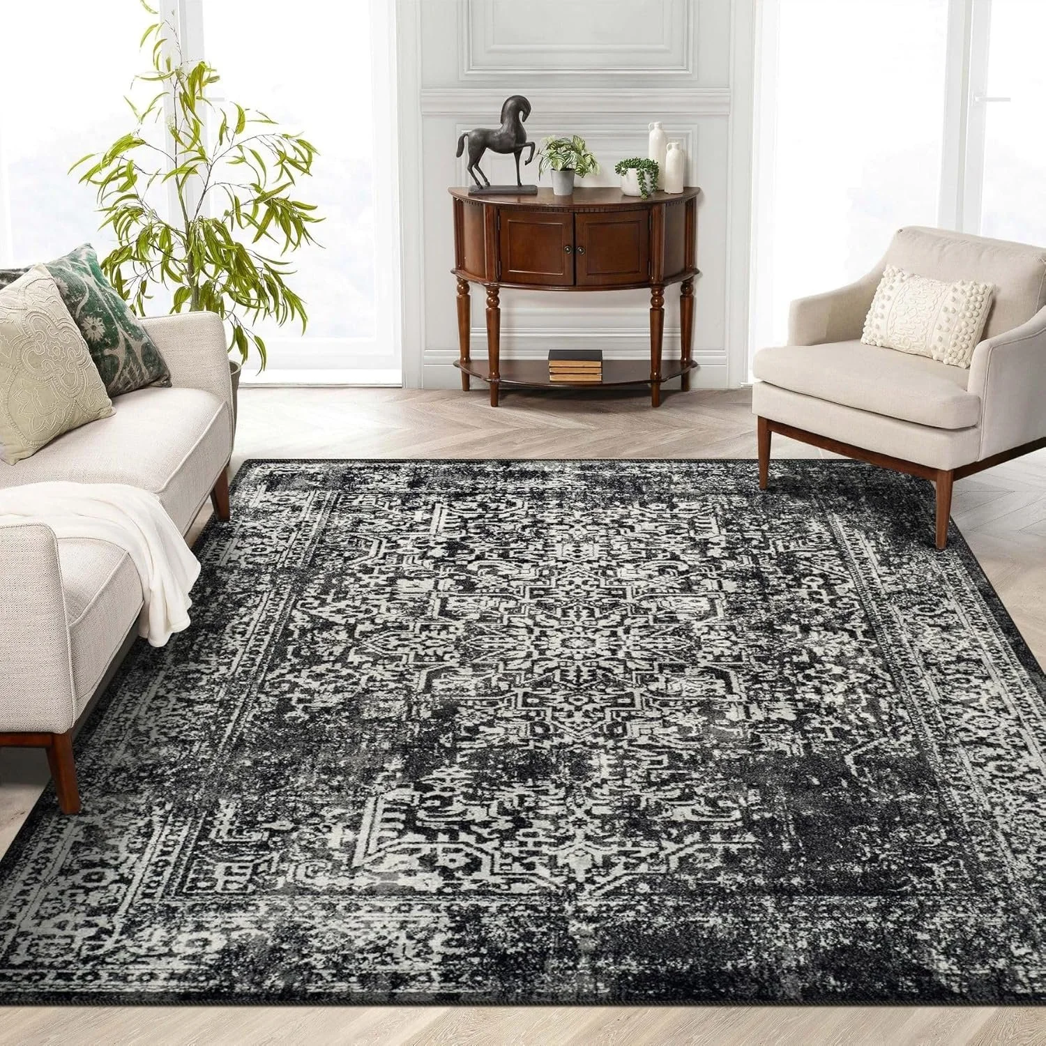 A living room with a black and white patterned area rug, a beige sofa with pillows, a white armchair with a pillow, a large green plant, and a wooden console table with decorative items against a white wall and light-colored wood flooring.