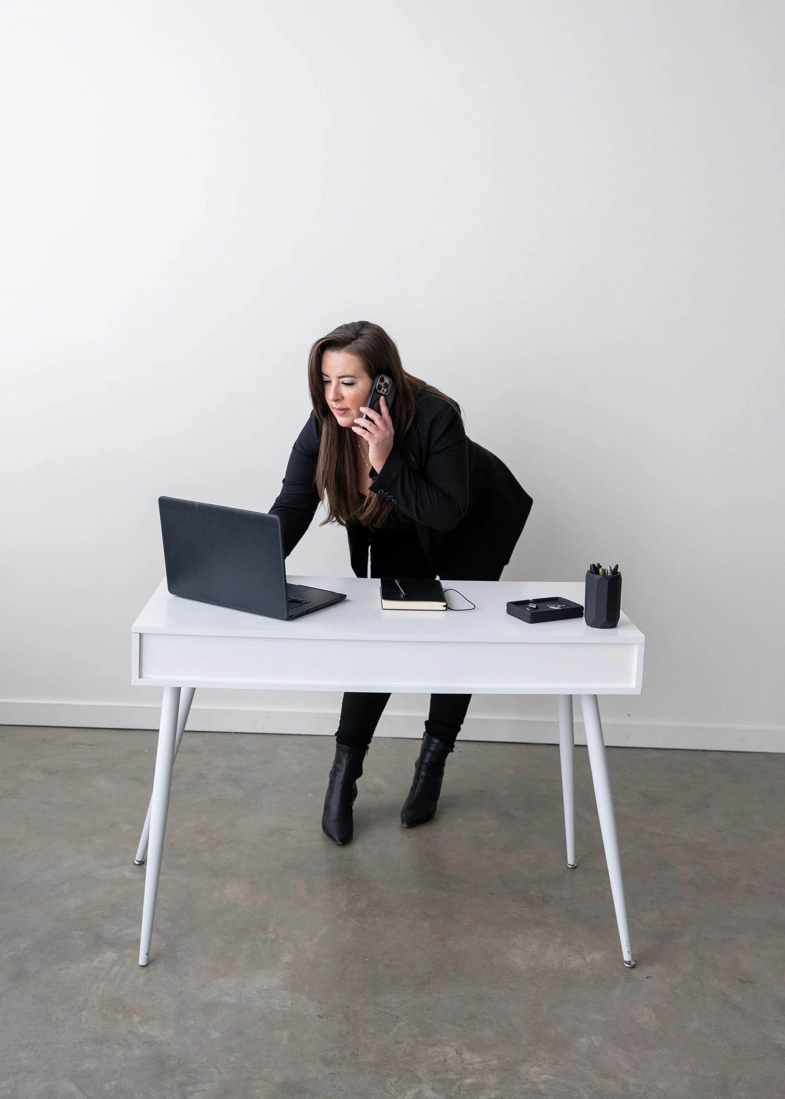 Kelly Abesdris, Founder of MTTE BLLK Agency, in a black outfit leaning over a white desk, talking on a cell phone, with a laptop, a notebook, a pen holder with pens, and a black case on the desk, against a plain white wall.