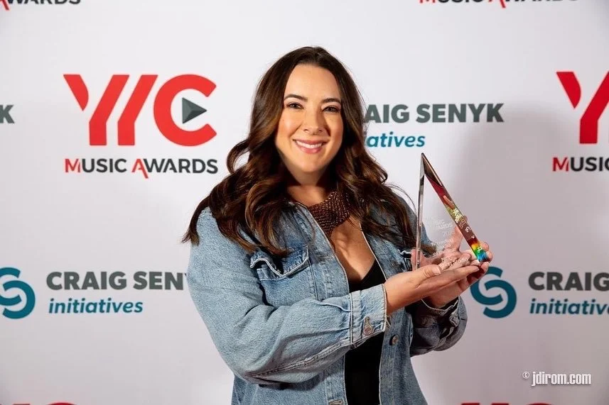 Kelly Besd wearing a denim jacket, smiling and holding a clear award trophy at the YYC Music Awards event, standing in front of a backdrop with event logos and sponsors.