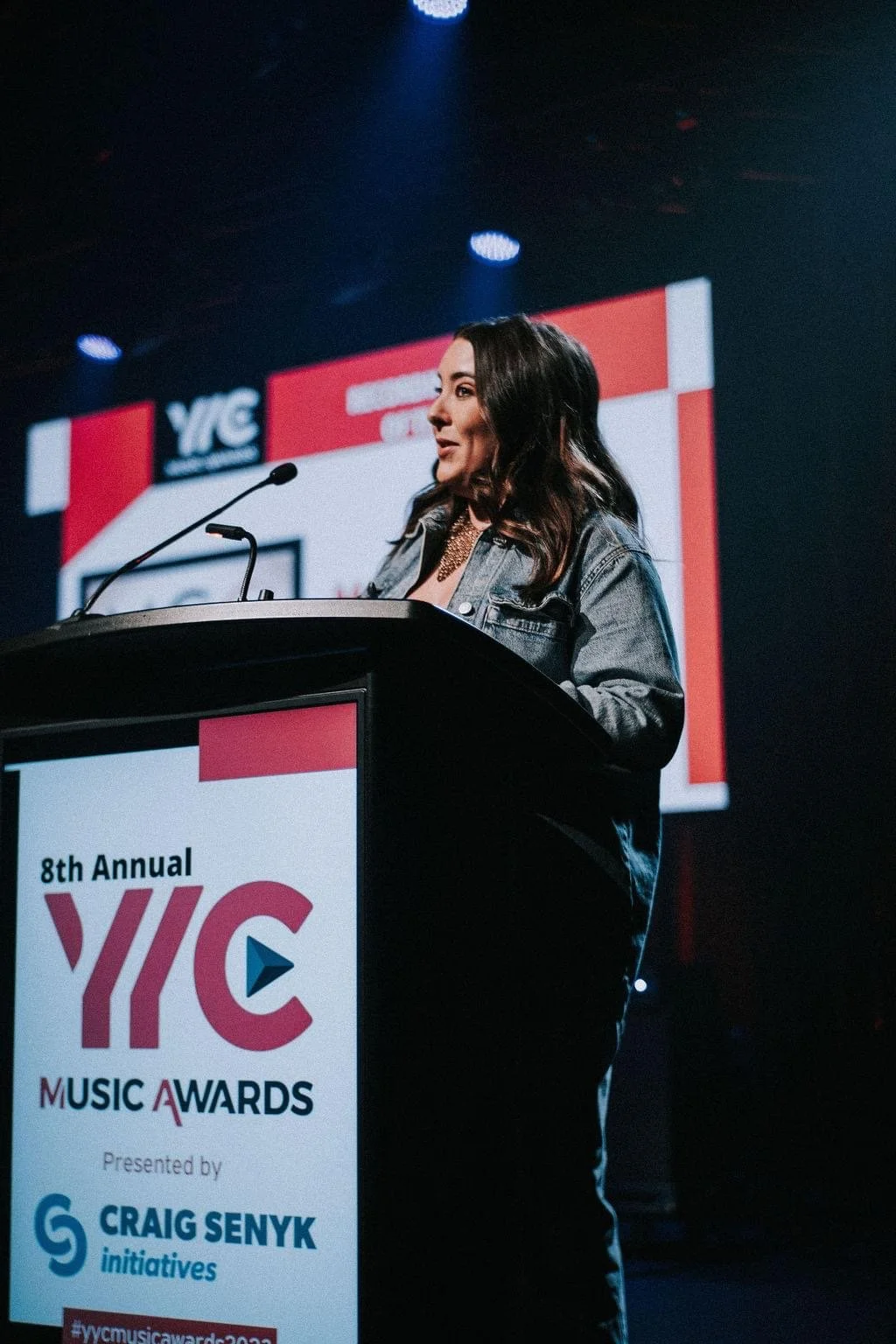 Kelly Besd accepting an award at a podium during the 8th Annual YYC Music Awards, with a large screen displaying the event's logo in the background.