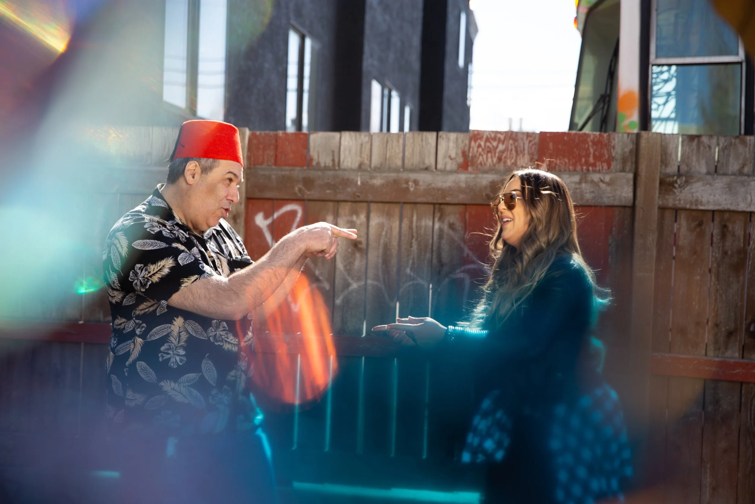 A man wearing a red fez hat and a black shirt with white leaf patterns points and talks with a woman wearing sunglasses and a dark jacket. They are outdoors behind a wooden fence with graffiti, with city buildings and windows in the background, and sunlight creating lens flares.
