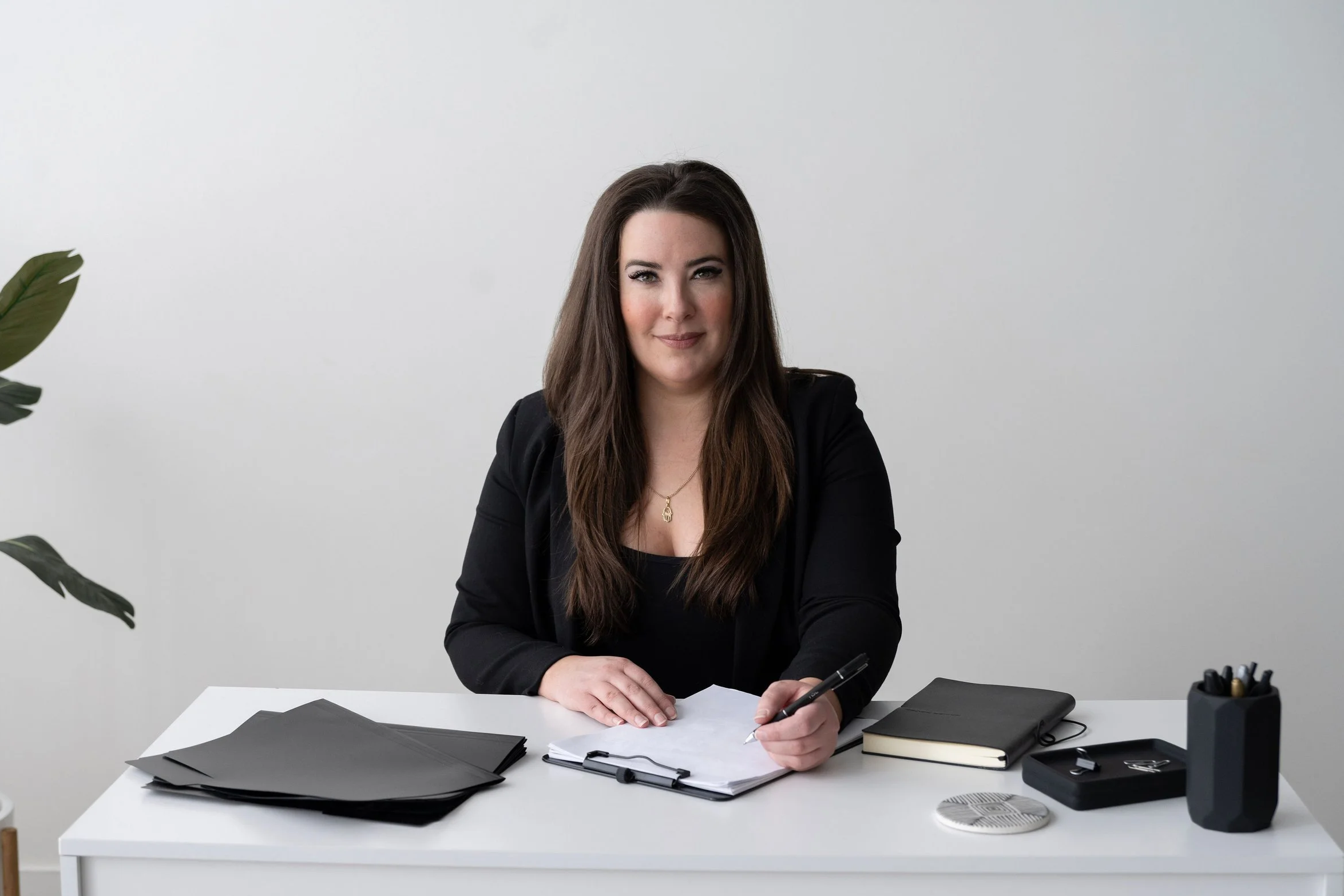 Kelly Abesdris, Founder of MTTE BLLK Agency, wearing a black blazer, sitting at a white desk with black folders, a notepad, and a pen, against a plain white wall, with some green leaves visible on the left side of the image.