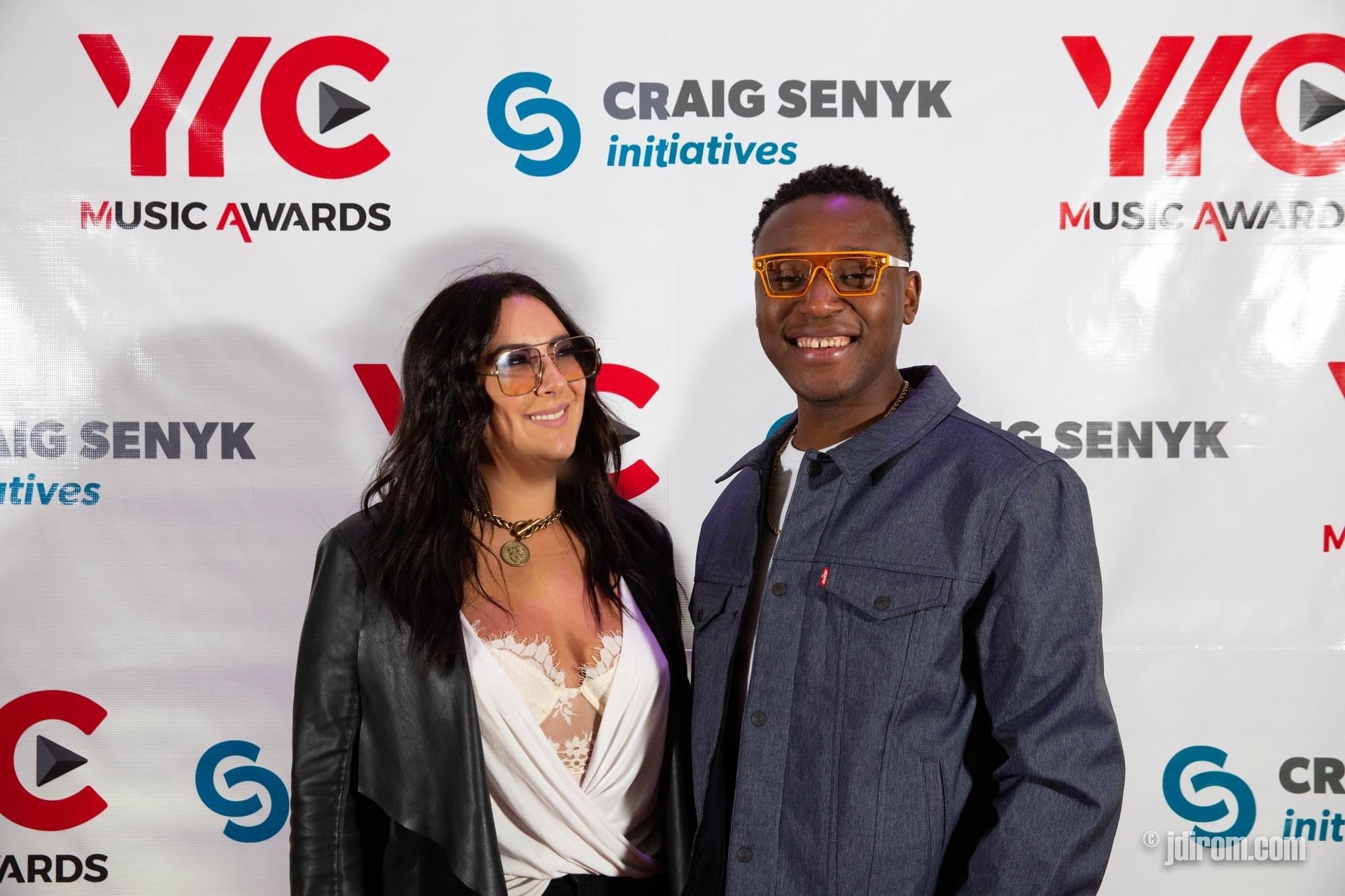 Two people posing at the Craig Senyk Initiatives and YVIC Music Awards event, standing in front of a backdrop with logos and text. The woman has long dark hair and is wearing glasses, a black leather jacket, and a white blouse with lace detailing. The man has short hair and is wearing large orange glasses and a denim shirt.