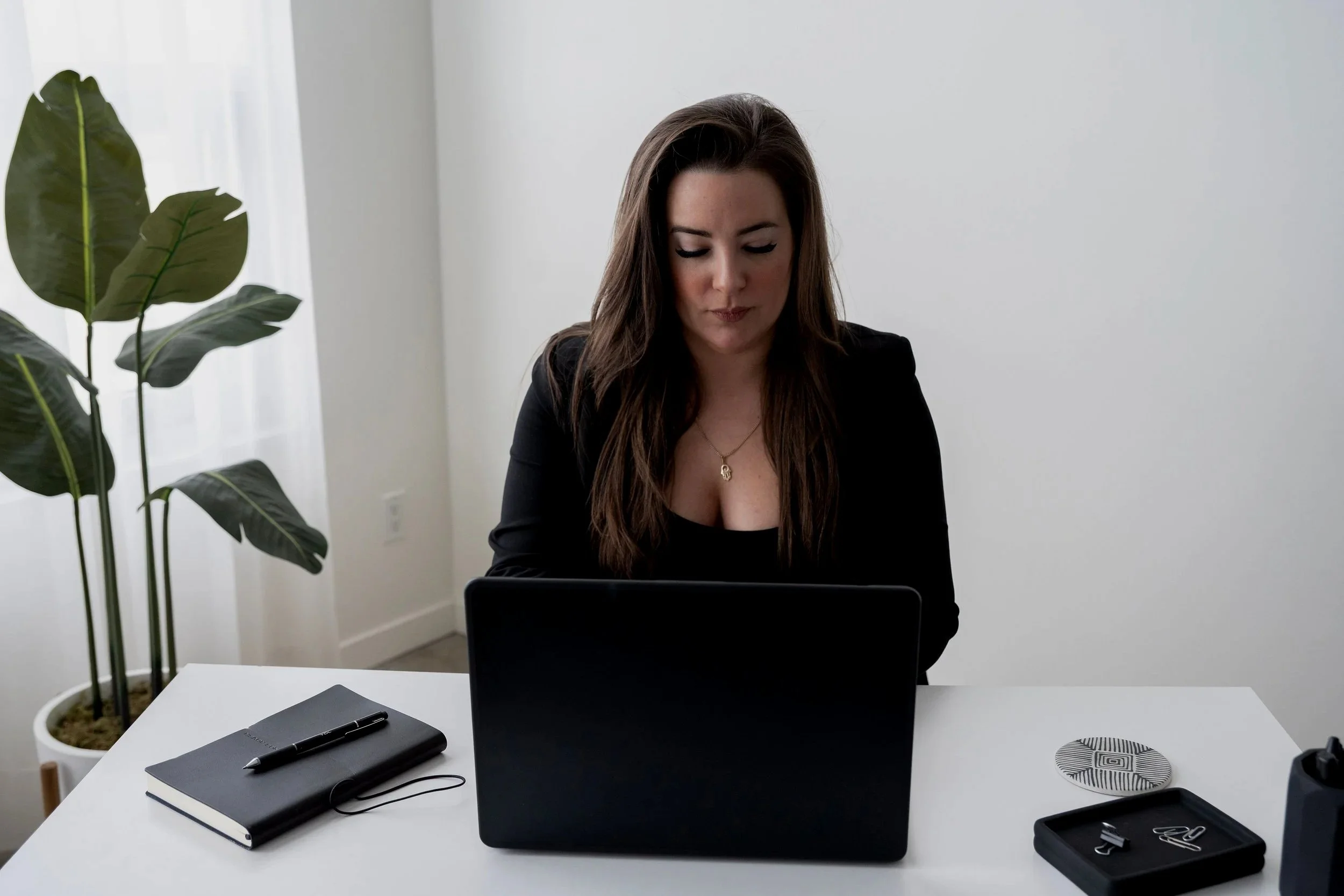 A woman with long brown hair looking down at her computer screen in a minimalist office. There is a potted plant on the left, a black notebook with a pen on top, and a wireless speaker on the desk. The background features a white wall and a large window with sheer curtains.