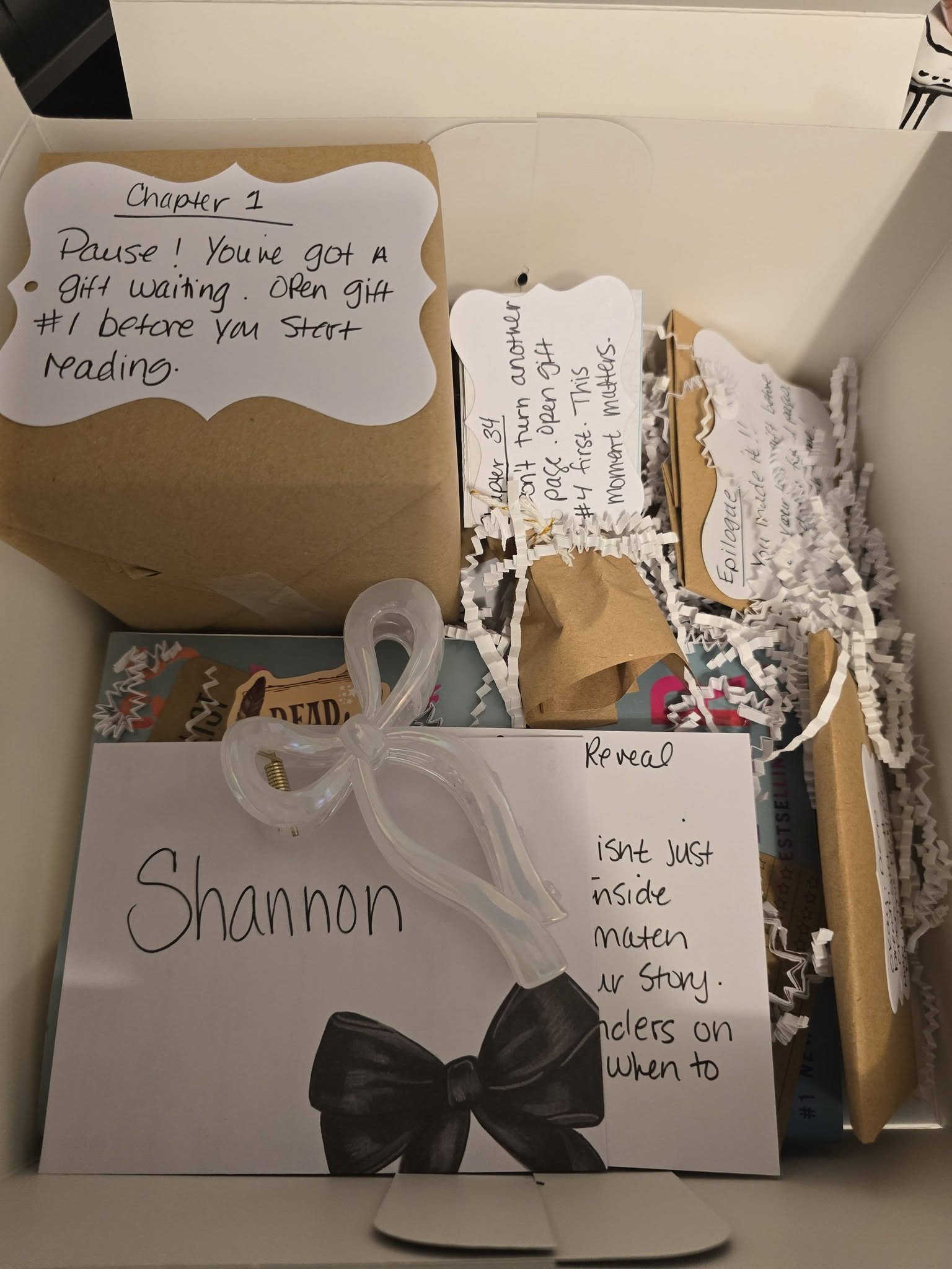 A white card with handwritten notes about a chapter in a book. The notes include instructions to open gift #1 before starting reading, and a comment about the story and its themes. There are decorative paper shredded around the card, a clear bow-shaped clip, and some partially visible books and papers in the background.
