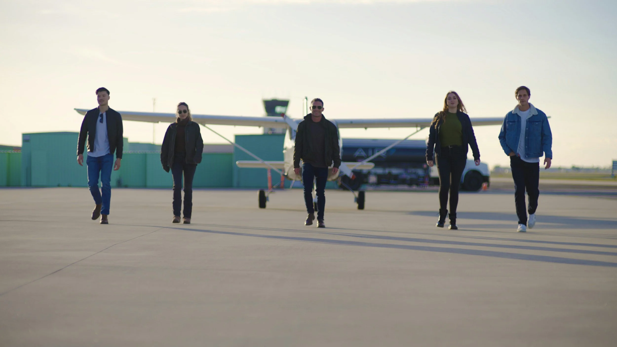 Group of five young adults walking on airport tarmac with small airplane and green hangar in background, sunny day.