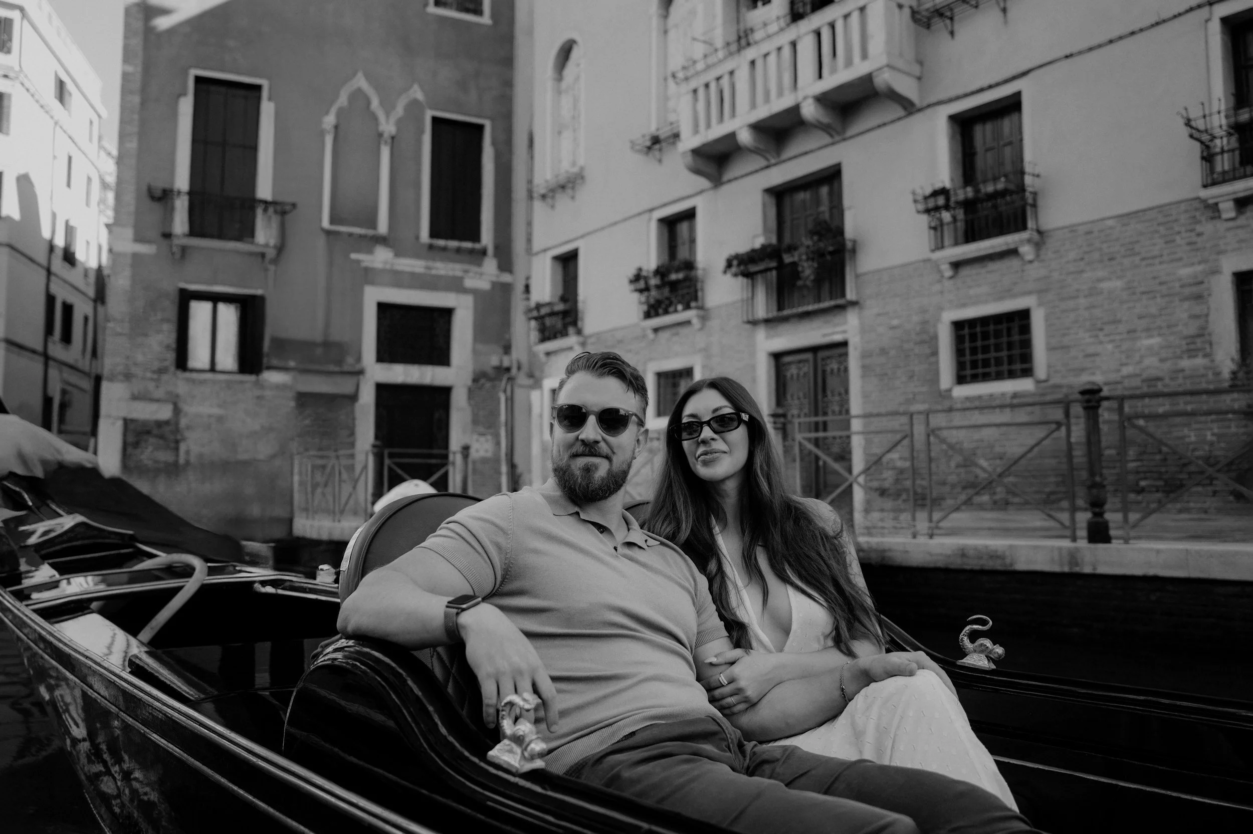 A couple with sunglasses sitting in a vintage car on a canal street in Venice, Italy, with historic buildings in the background.
