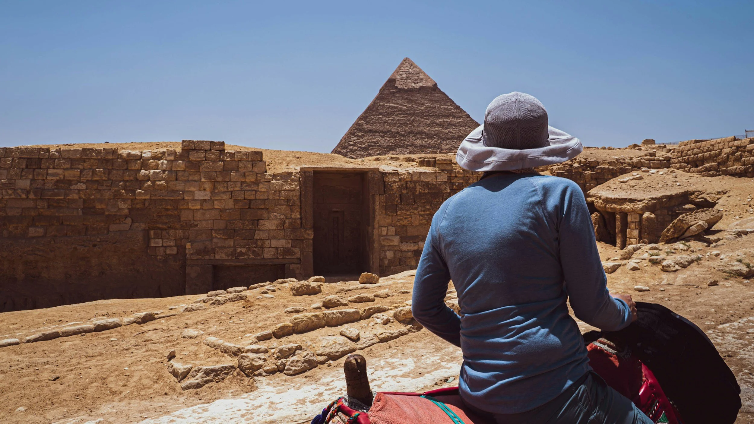 A person wearing a wide-brimmed hat and a blue long-sleeve shirt sitting on the ground, looking towards the Pyramid of Khafre in Giza, Egypt, with ancient ruins and a blue sky in the background.