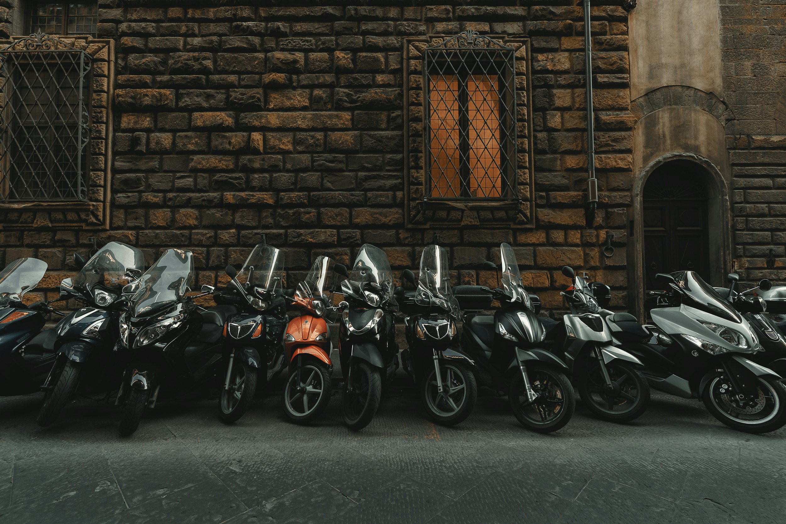 Row of parked motorcycles in front of a brick building with barred windows and a wooden door.   Throttle Therapy trip vibes.