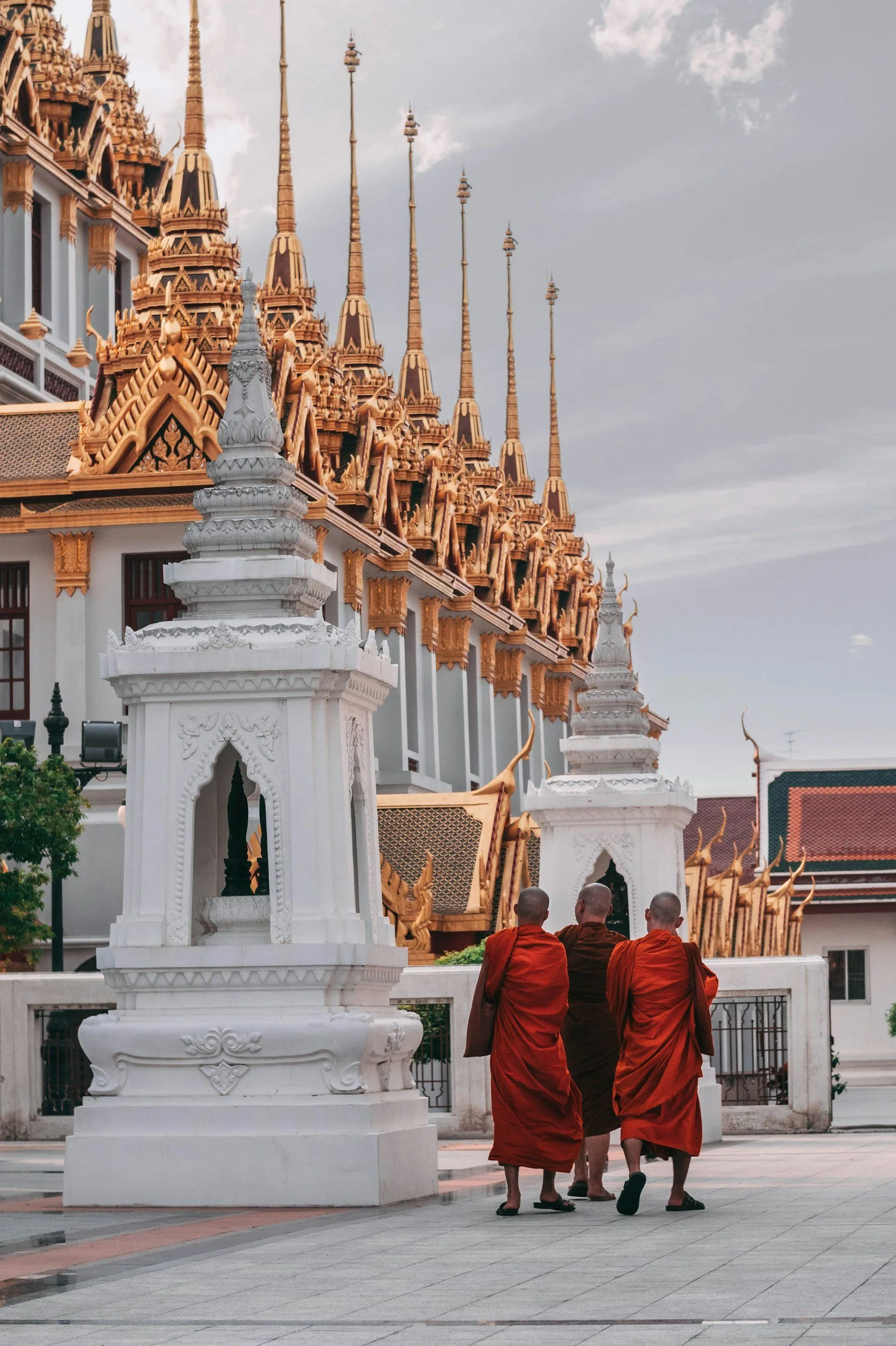 Three Buddhist monks in orange and brown robes walking together in a temple courtyard with ornate temple architecture and white decorative structures.