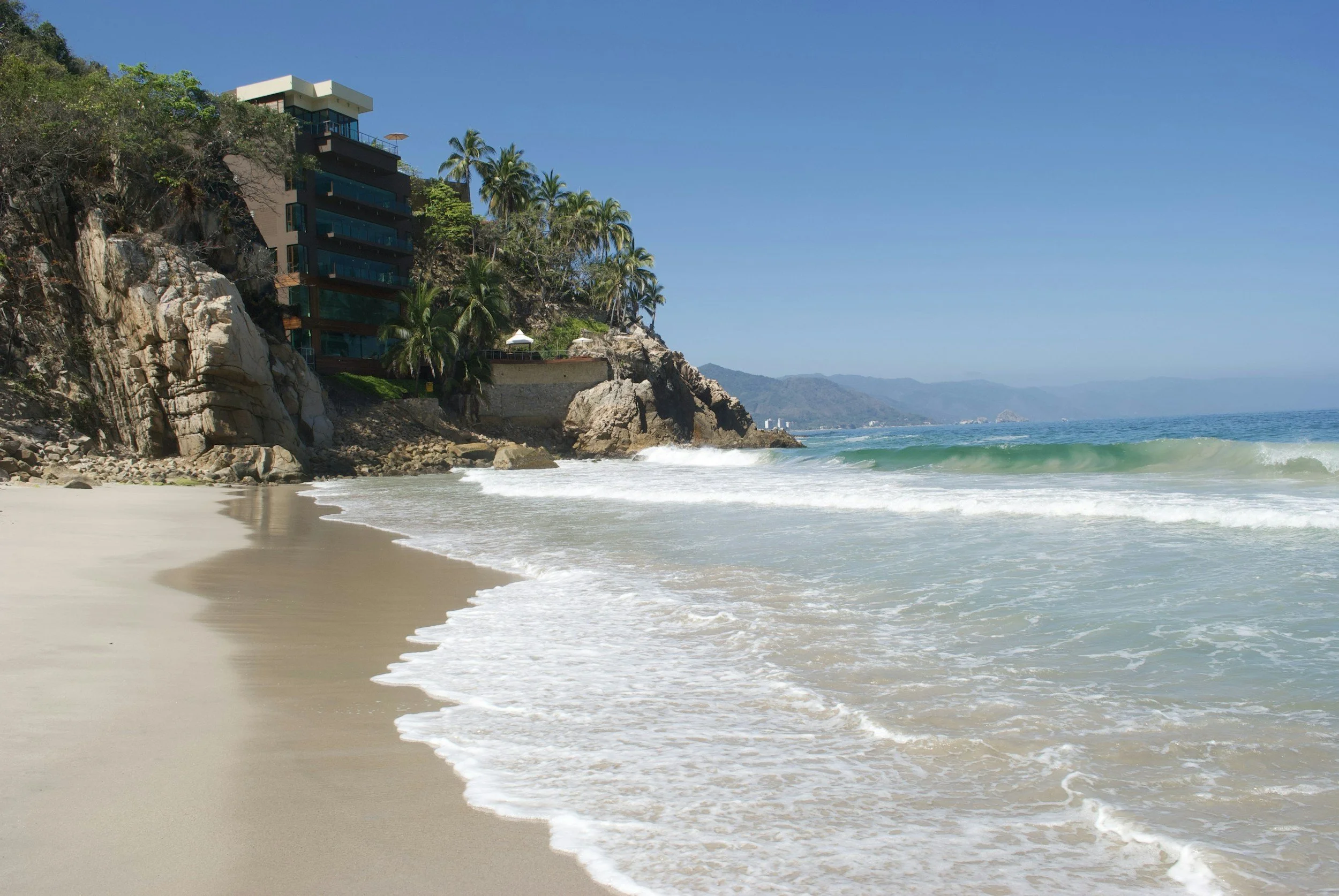 A beach with white sand and gentle waves, with a rocky hillside featuring a modern building, palm trees, and umbrella shades, overlooking the ocean under a clear blue sky.