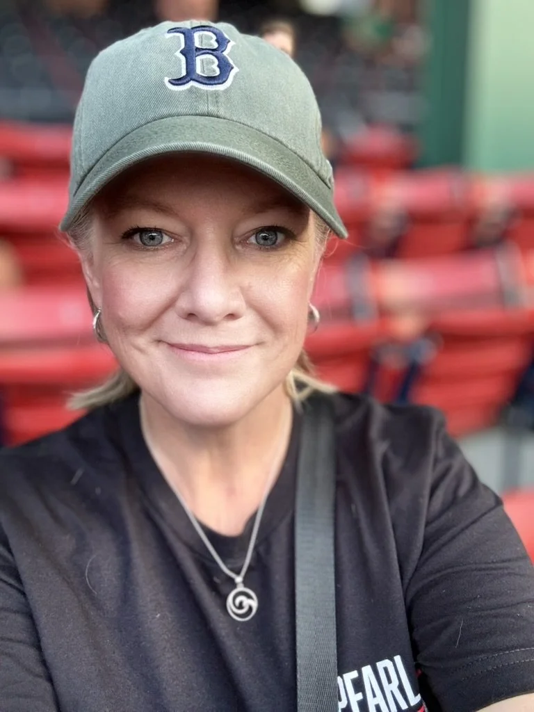 A woman with blonde hair wearing a gray baseball cap with a blue 'B' logo, black shirt, silver necklace with a swirl pendant, and silver hoop earrings, taking a selfie at a stadium with red seats in the background.