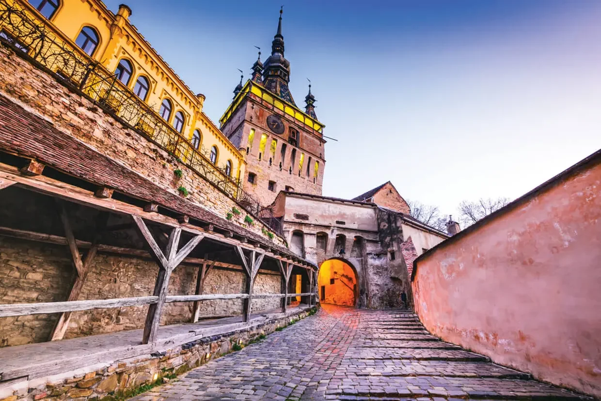 A cobblestone walkway leads through an arched stone entrance with a tall, ornate clock tower in the background. The tower has a dark, pointed roof and multiple smaller spires, set against a clear sky with a gradient from blue to purple at sunset.